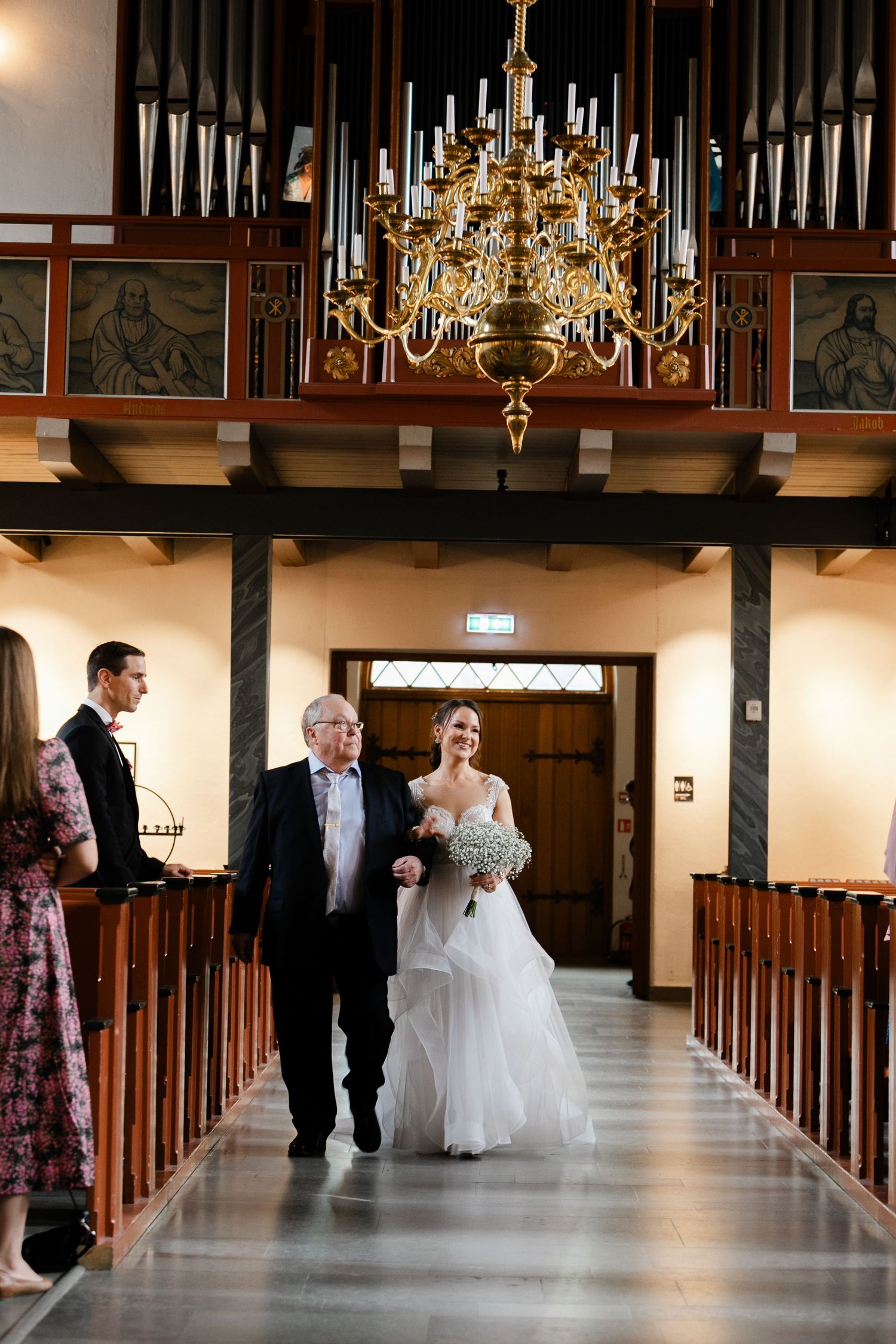 Wedding party walking down the aisle of a historic church with ornate chandeliers and wooden beams.