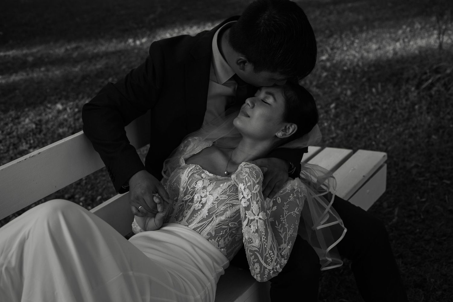 A romantic black and white wedding photo of a couple sharing an intimate moment on a bench in a garden.