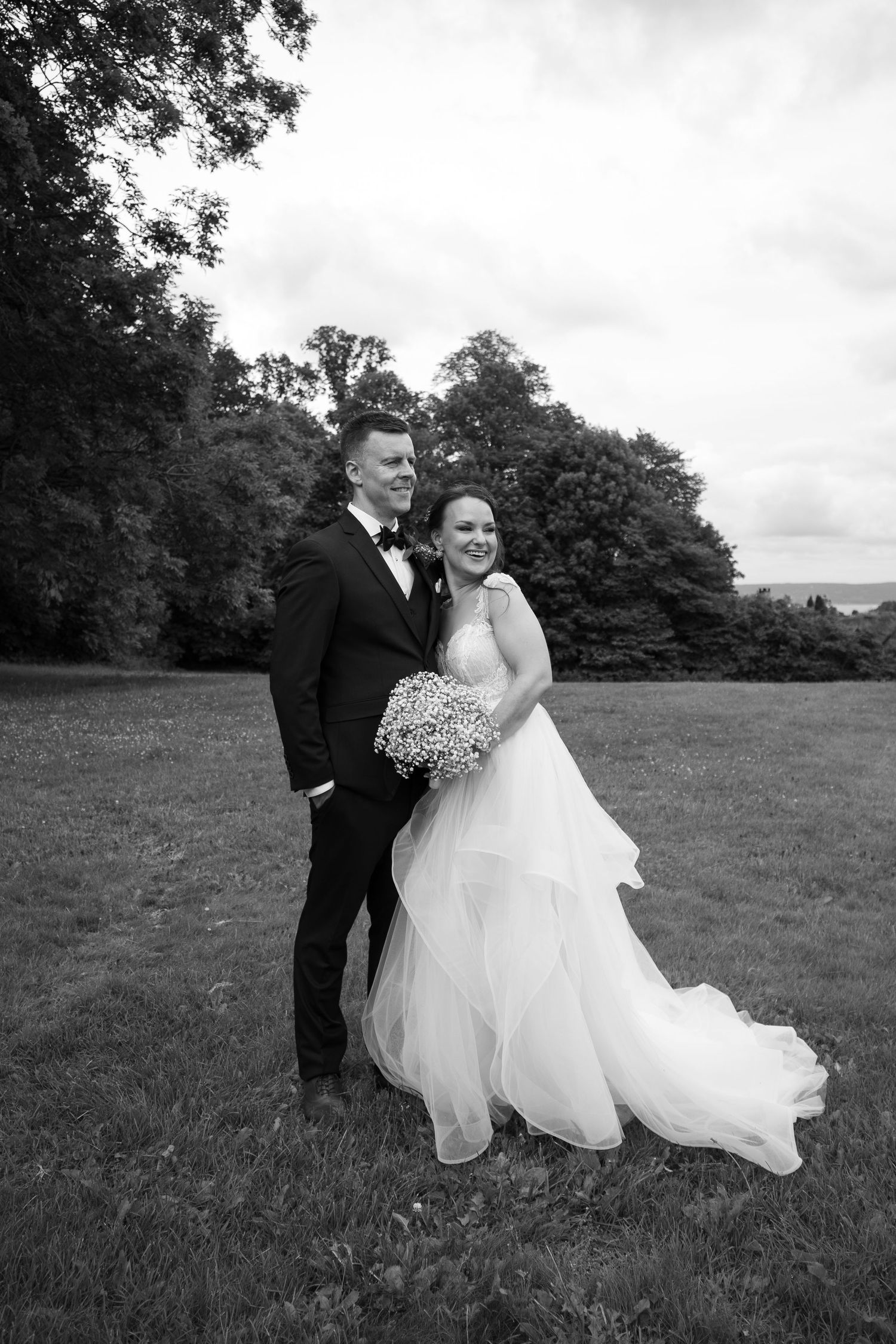 A couple in formal wedding attire walk together across a grassy meadow with trees in the background.