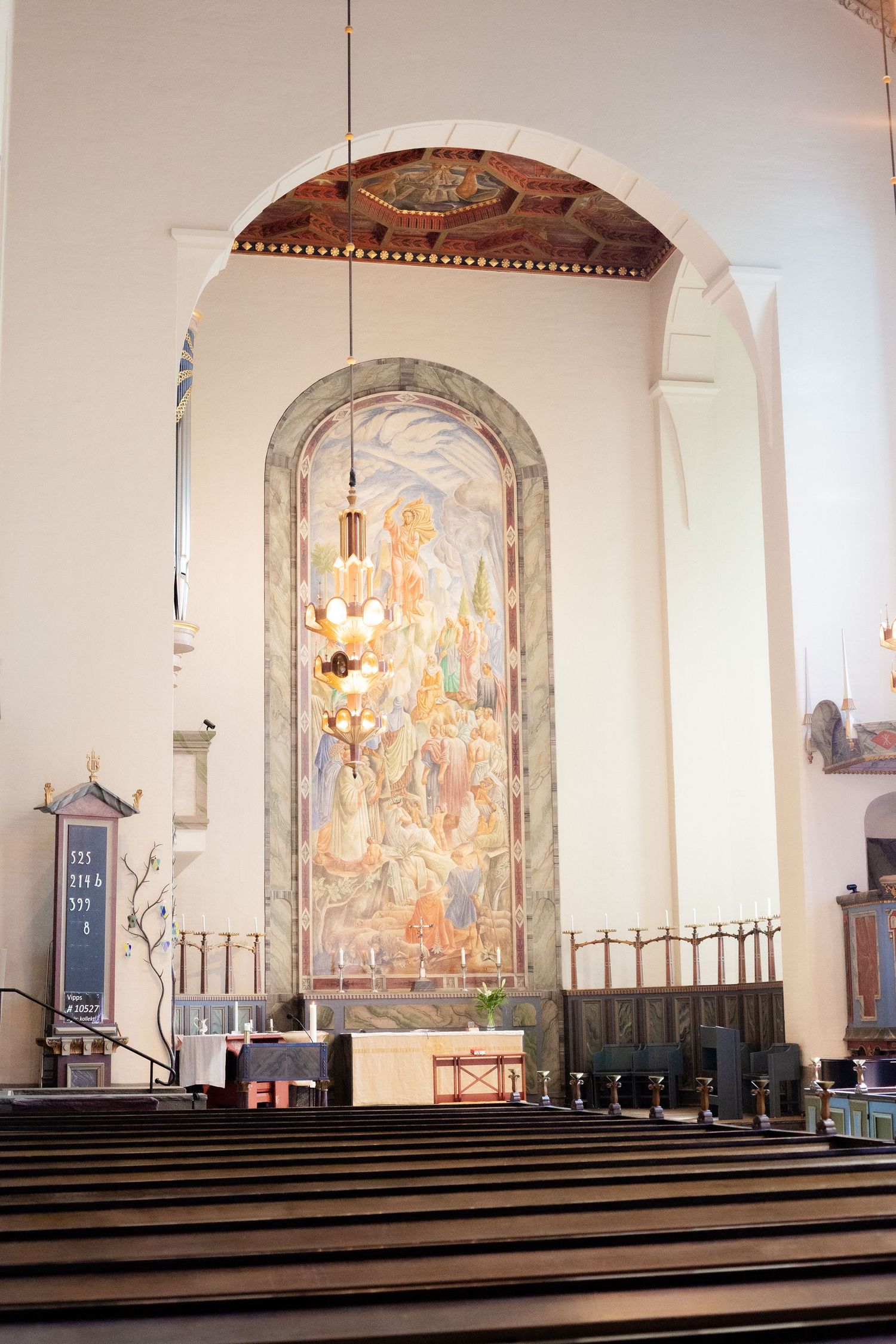 Interior of a church sanctuary with a large illuminated mural and chandelier under an arched ceiling.