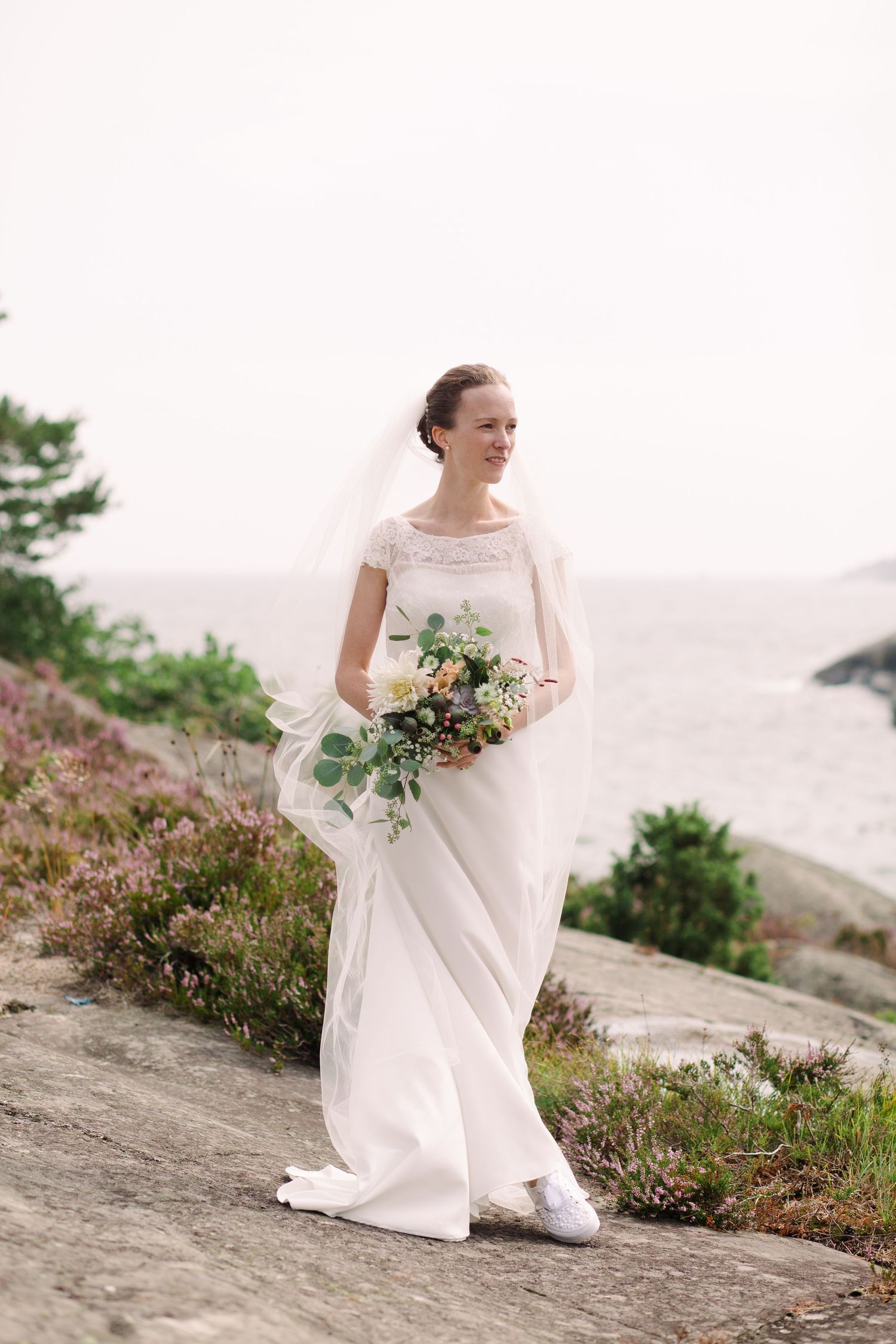 Bride in white dress holding bouquet on rocky coastal landscape.