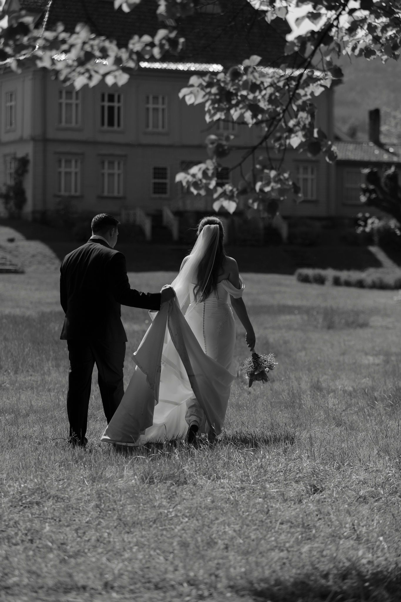 Black and white photo of bride and groom walking hand in hand across a grassy lawn toward an elegant manor house.
