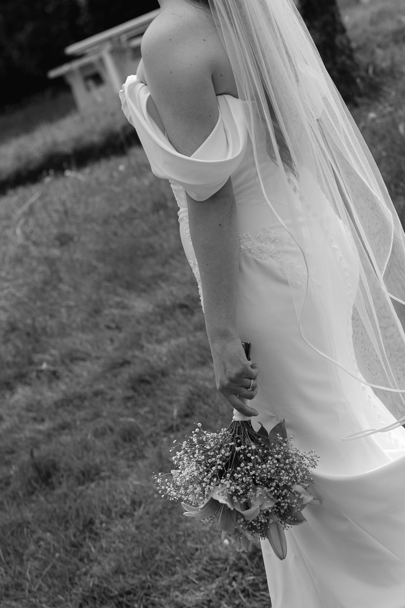 Black and white photo of a bride in an off-shoulder gown holding a bouquet, standing on grass with veil flowing.