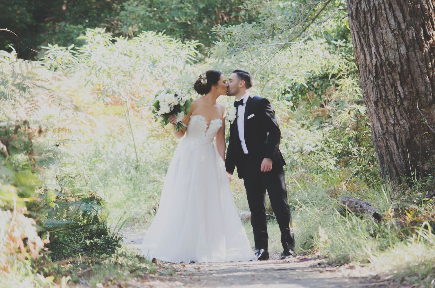A bride and groom share a romantic kiss on a sunlit forest path, with the bride in a white gown holding a bouquet.
