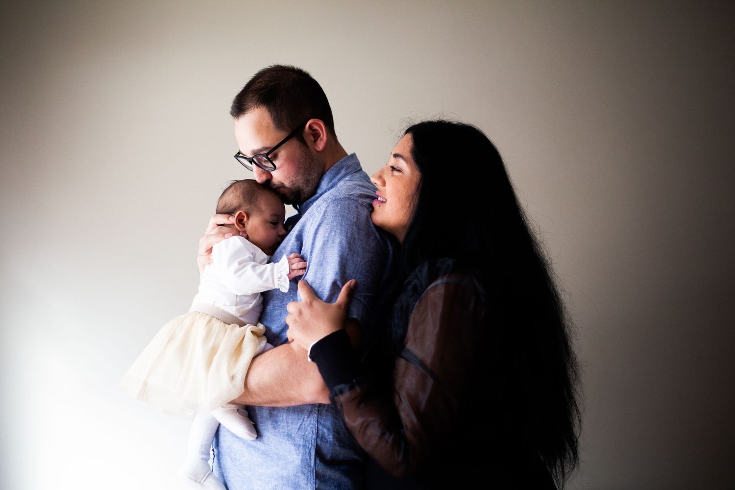 A family embraces in a tender moment against a simple background with dramatic lighting and shadows.