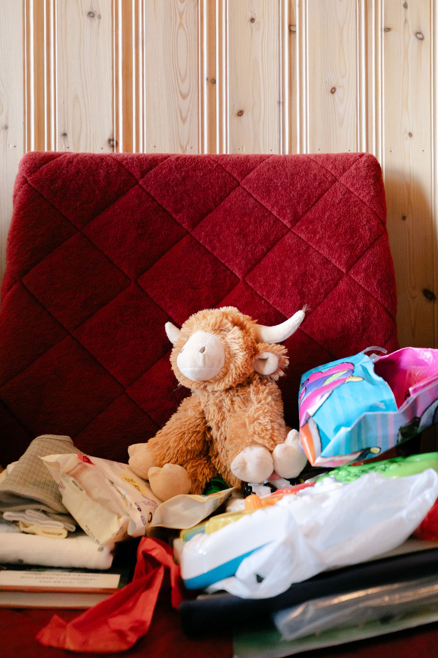 Stuffed animals and clothing items arranged on a red cushioned bench against wooden paneled walls.