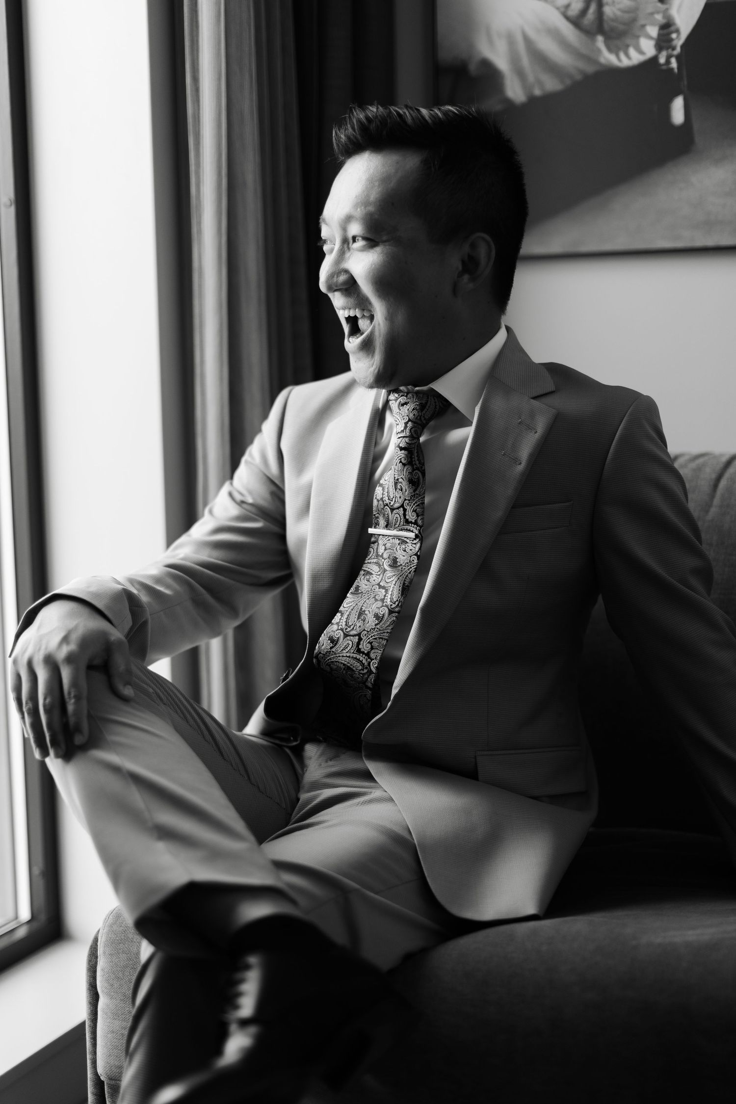 Black and white photo of groomsmen in suits lounging on chairs by a window.