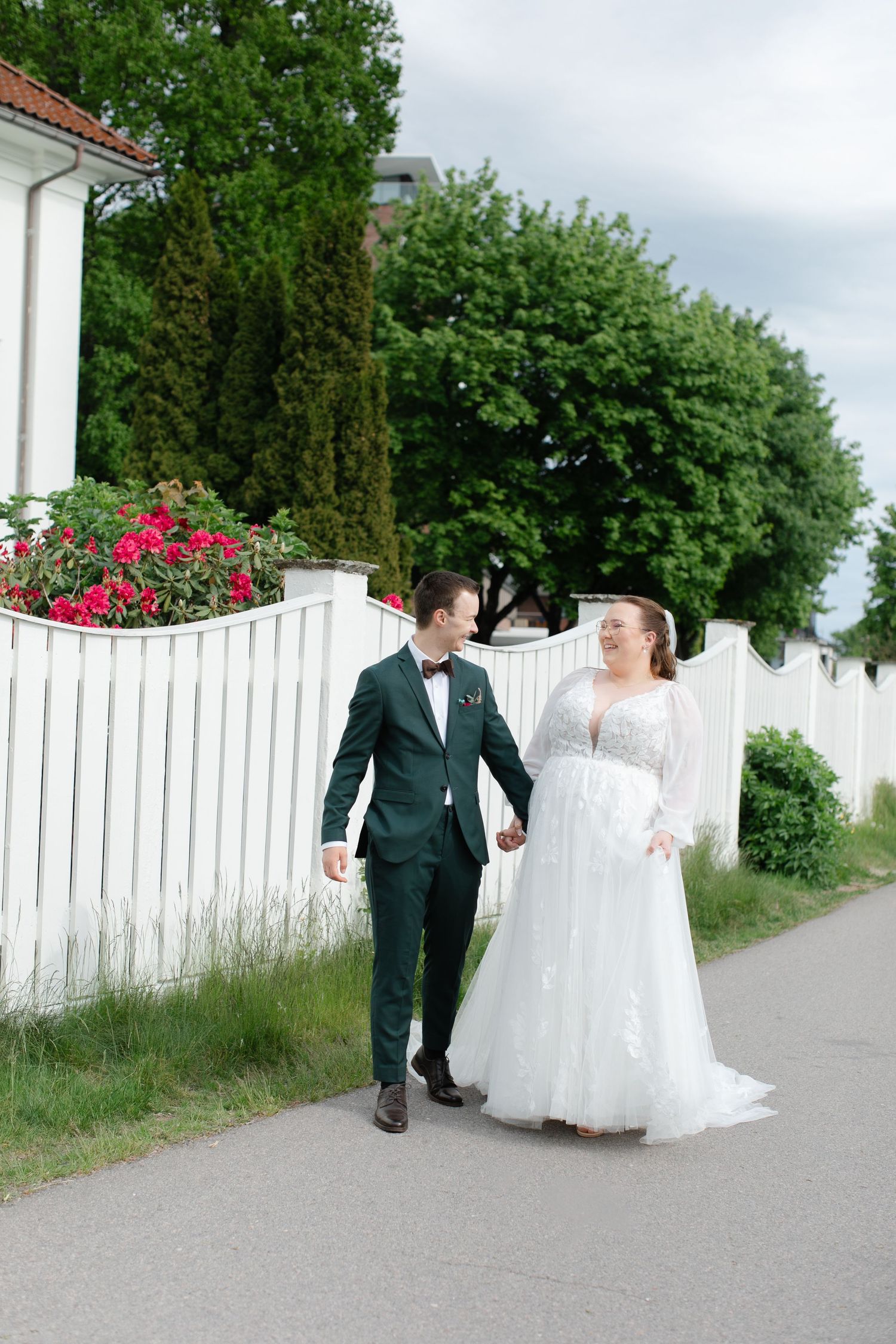 A bride and groom walk hand in hand along a path beside a white picket fence, surrounded by lush greenery.