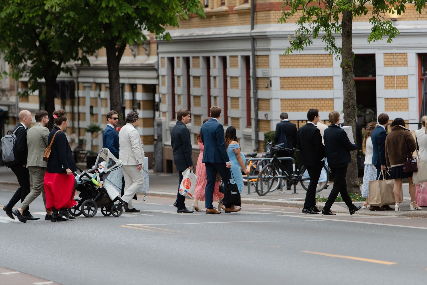 Crowd of people in formal attire crossing a busy urban street, with a baby stroller visible among the pedestrians.