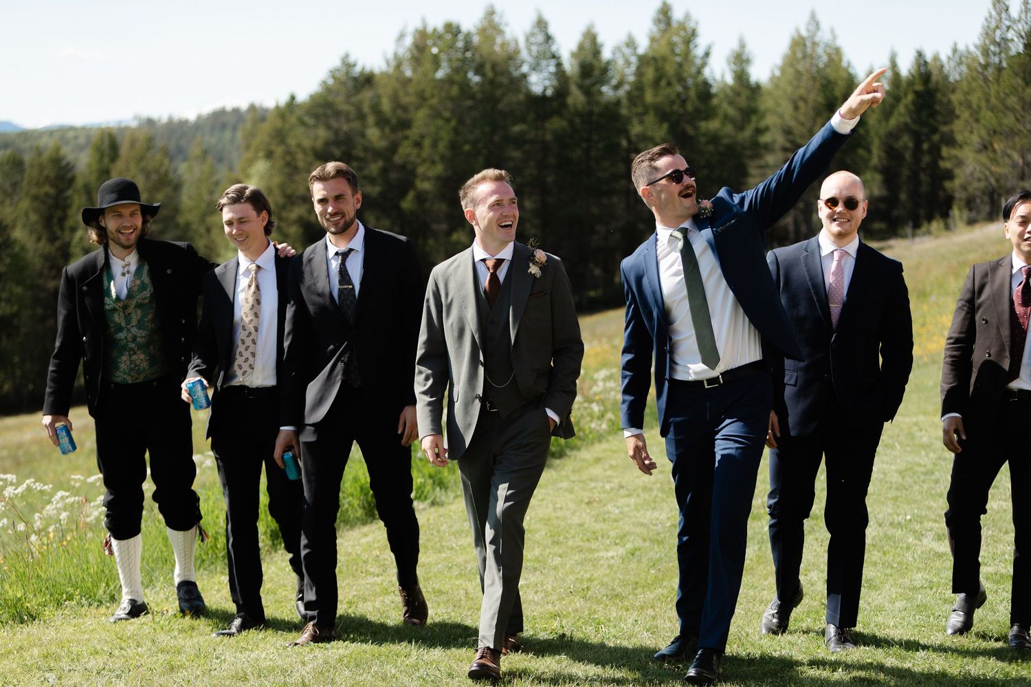 Groomsmen walking in a line through a grassy field with pine trees in the background.