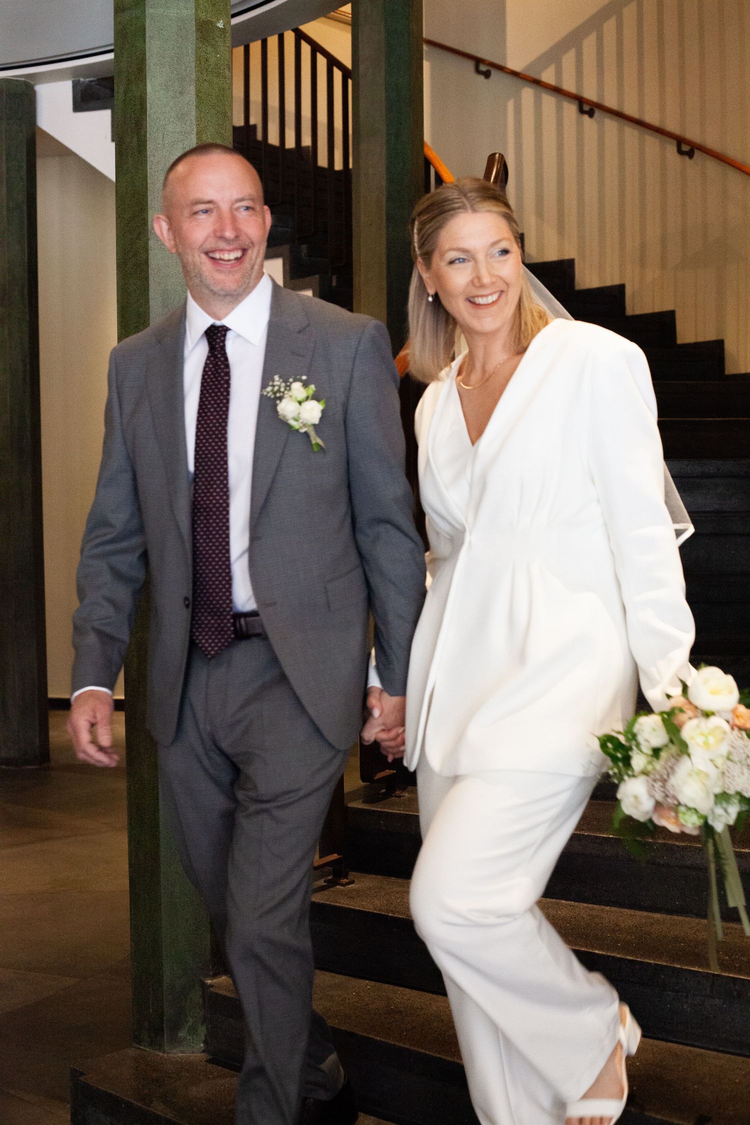 A couple walking together indoors, with the bride wearing a white dress and groom in a dark suit and burgundy tie.