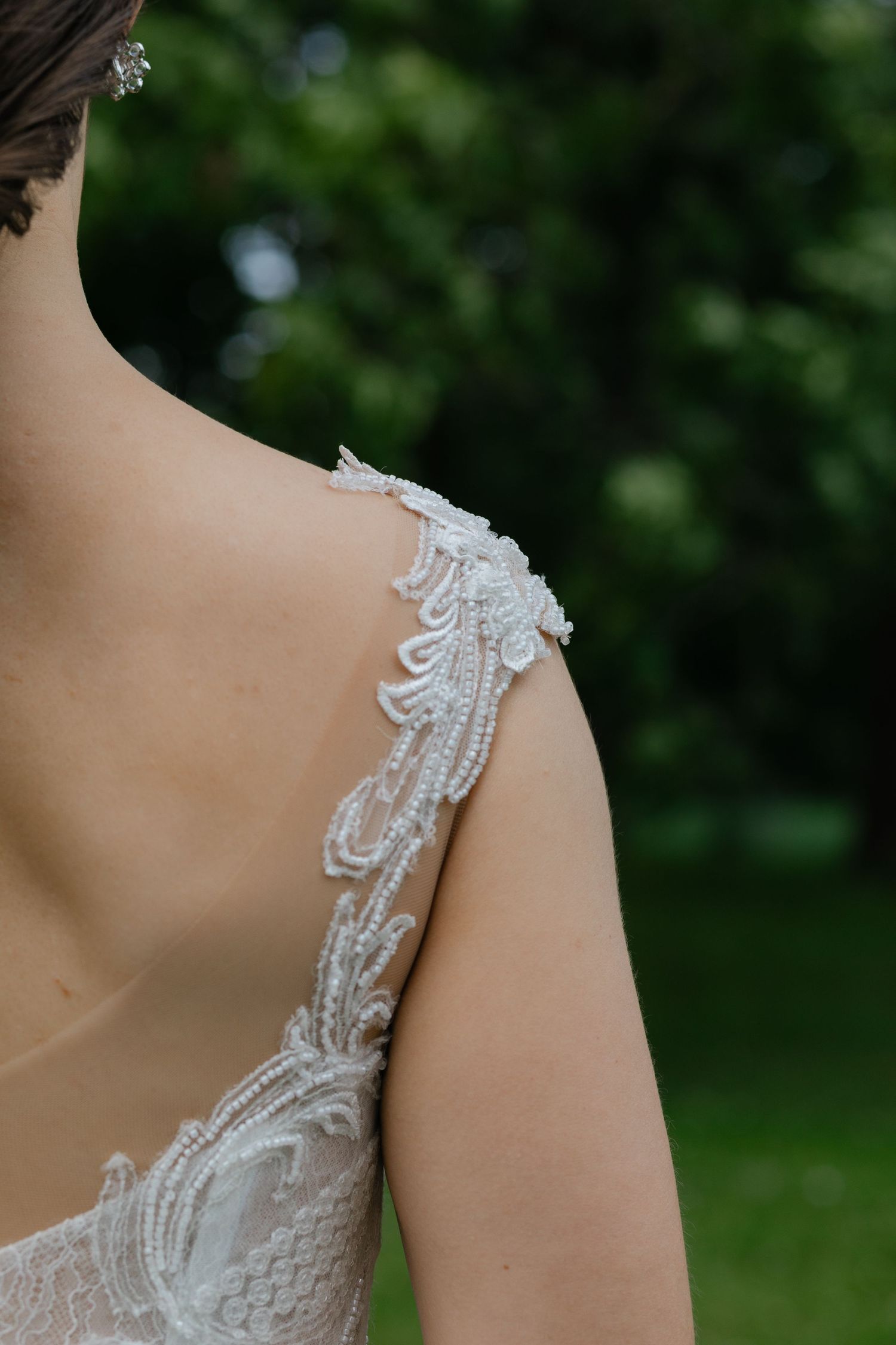 Close up of delicate beaded lace detail on wedding dress shoulder strap against green background.