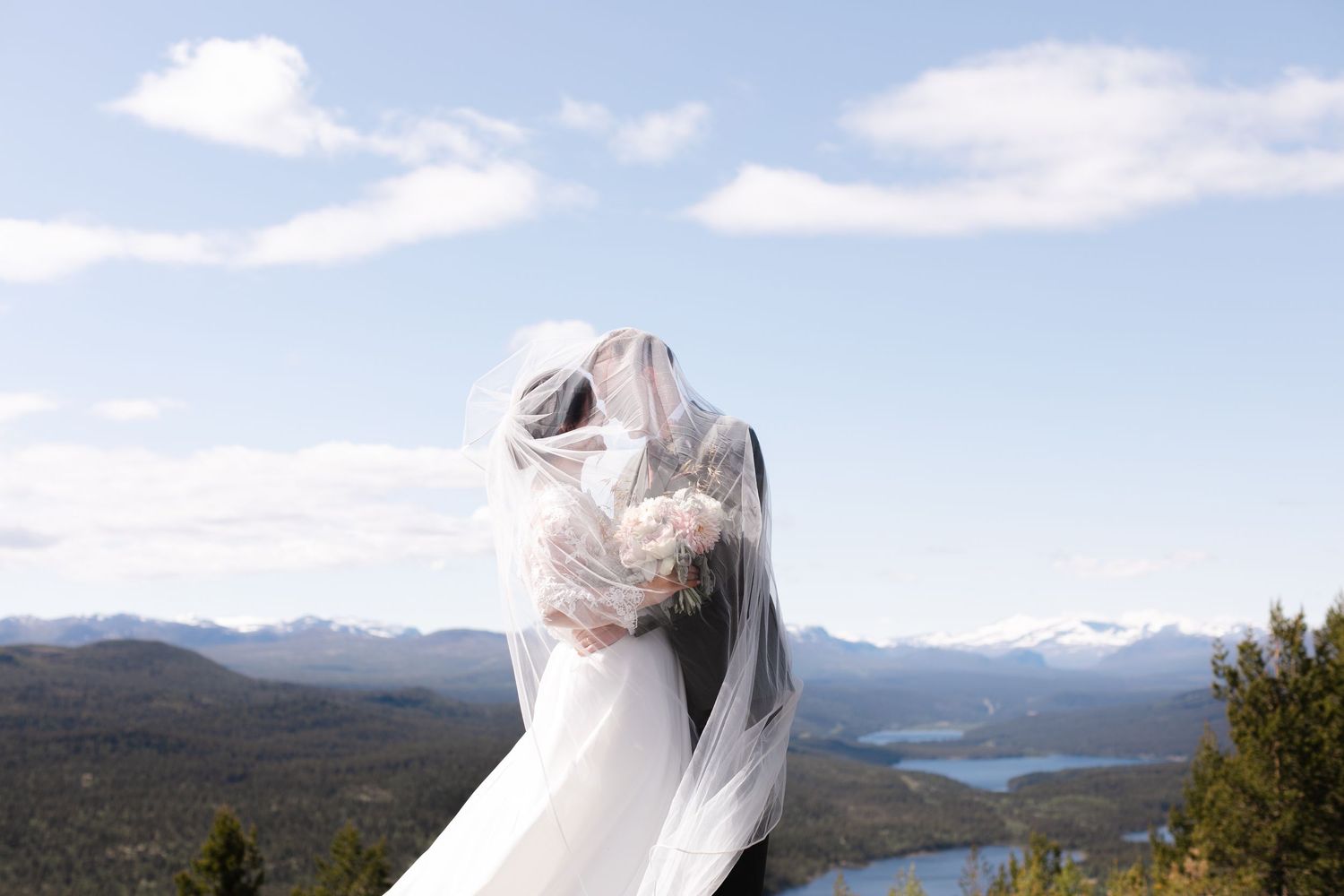 A couple in wedding attire embraces while overlooking a scenic mountain landscape with a lake in the distance.