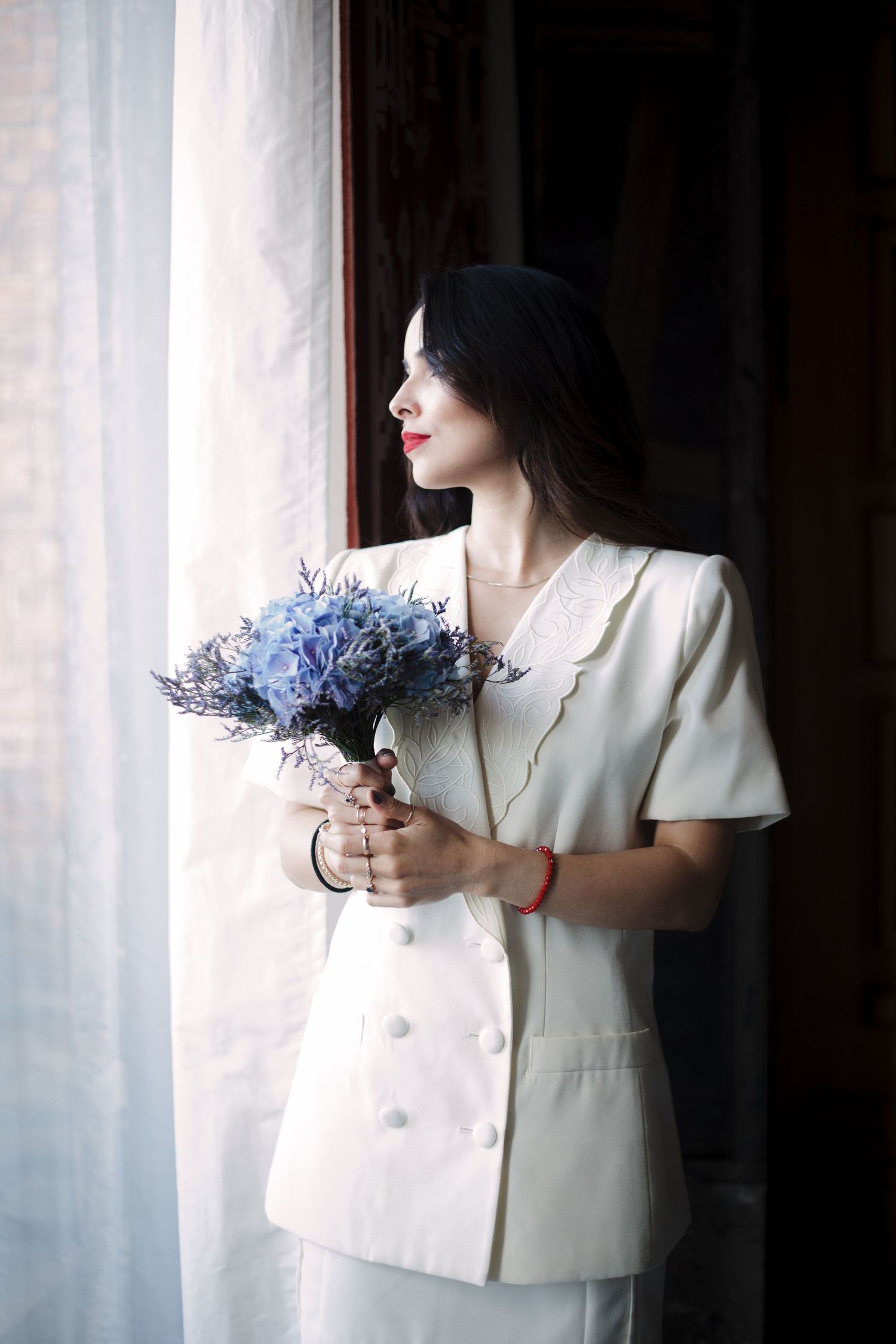 Person in white jacket holding blue flowers against soft window light.