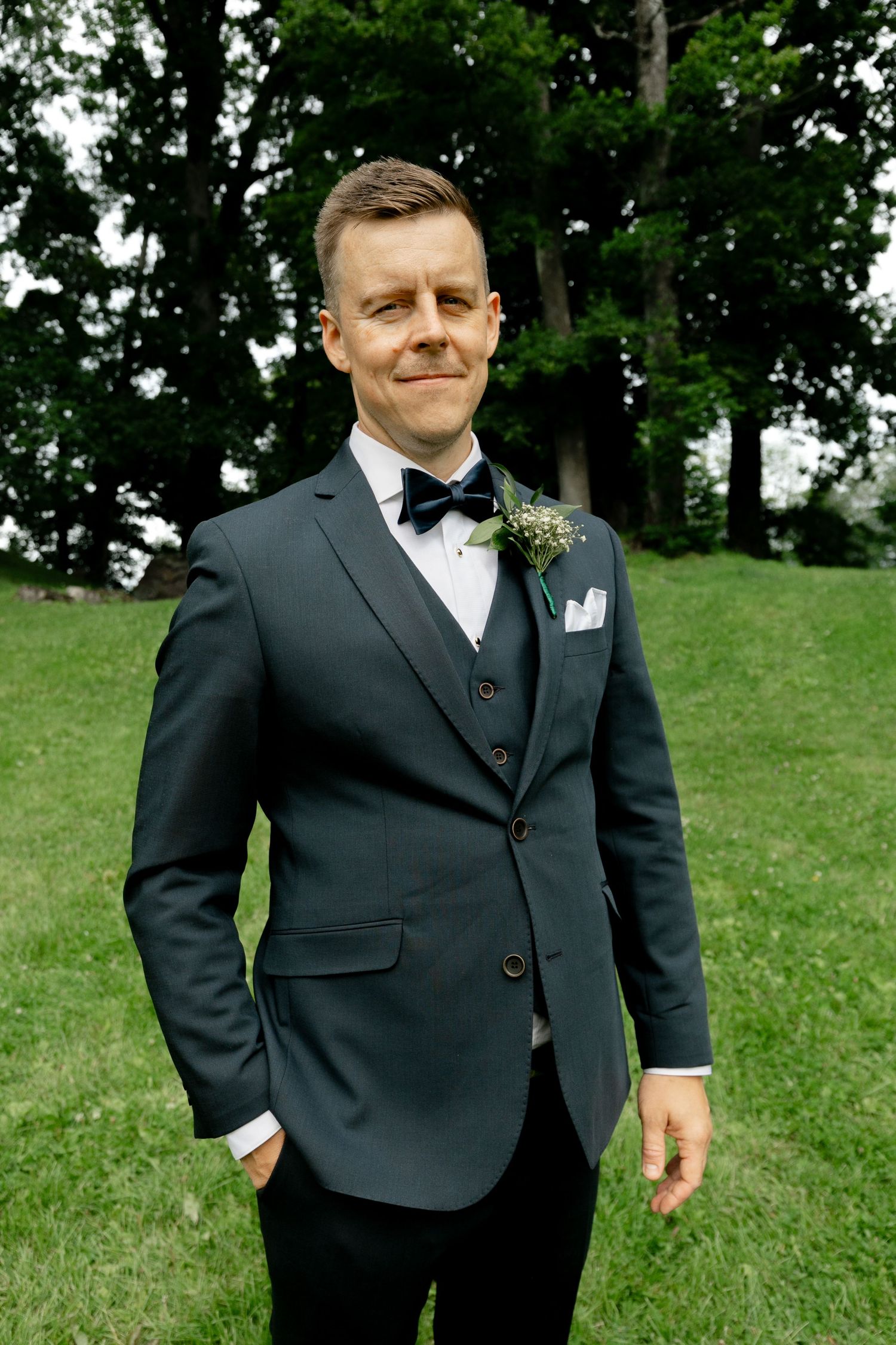Groom in black suit adjusts his tie while standing on grassy lawn with trees in background.