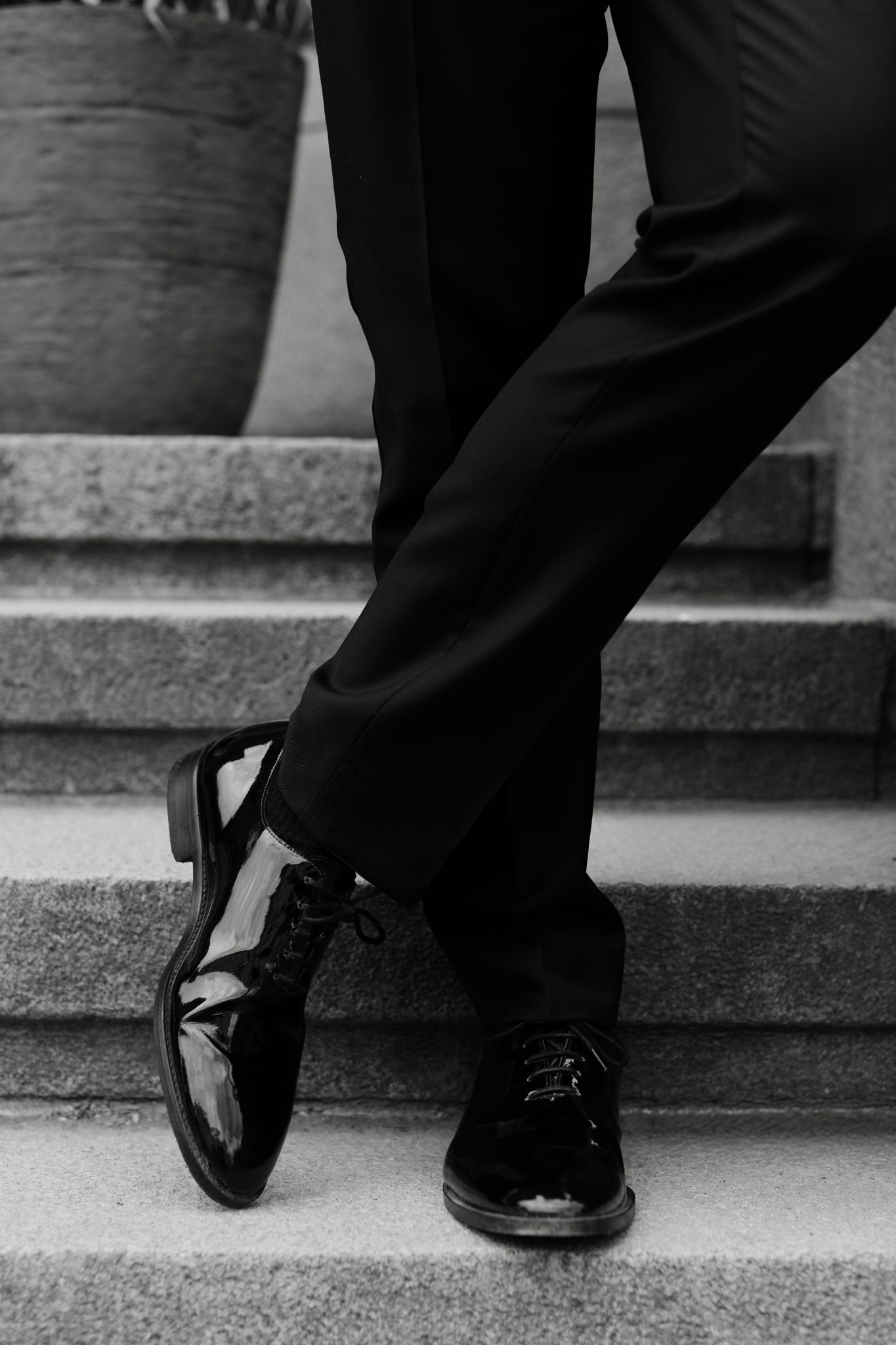 Black and white close-up of a groom wearing shiny patent leather dress shoes and black tuxedo trousers on stone steps.
