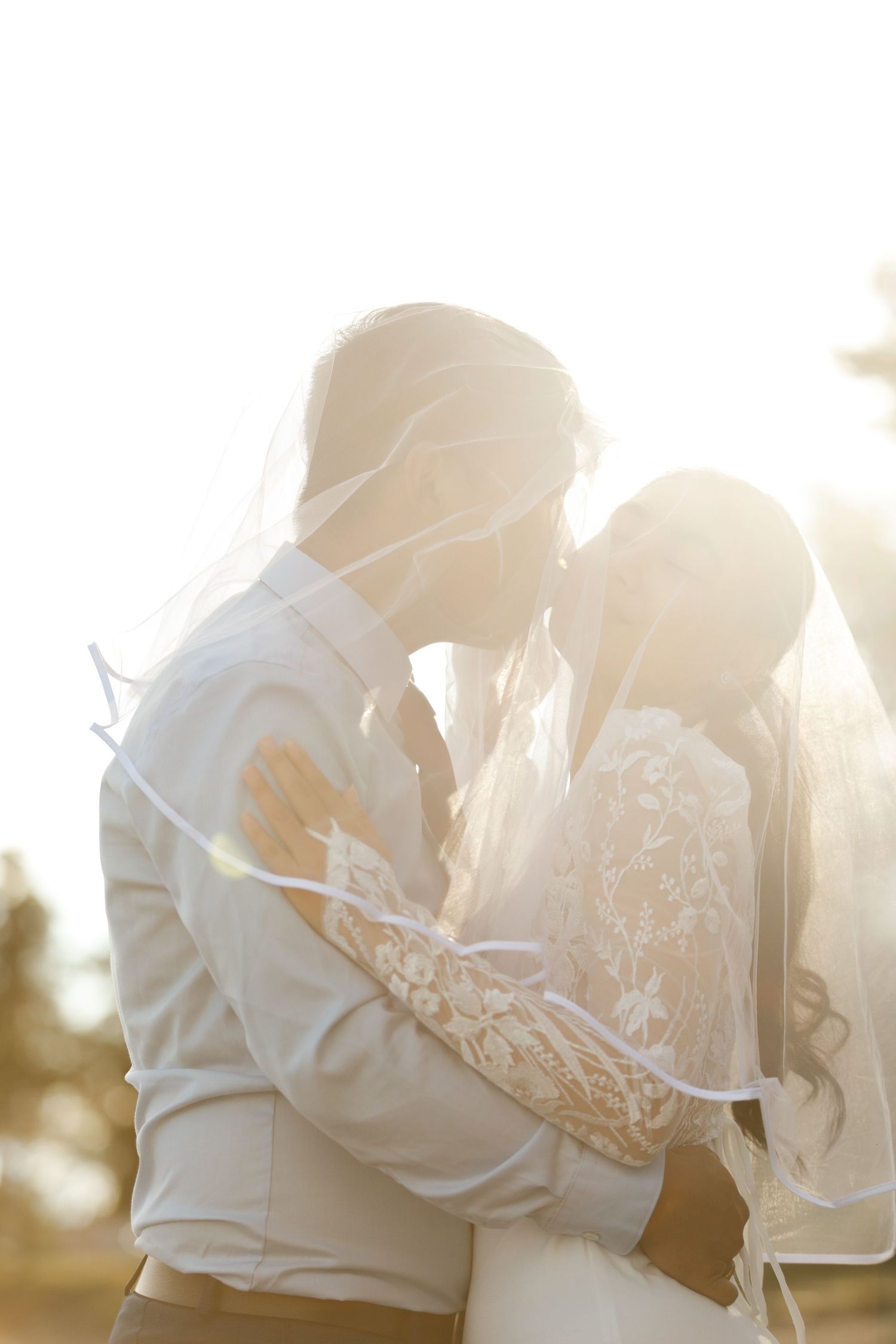 Bride and groom sharing a romantic kiss under a veil in golden backlit sunlight on their wedding day.