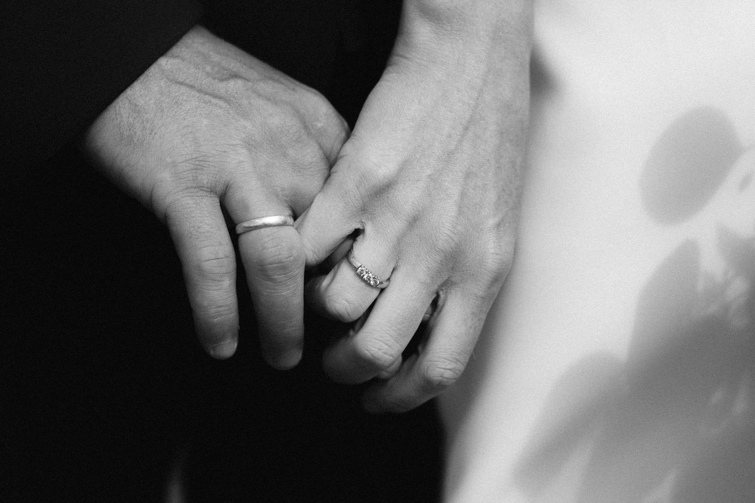 A black and white close-up photograph of two hands with wedding rings holding each other.