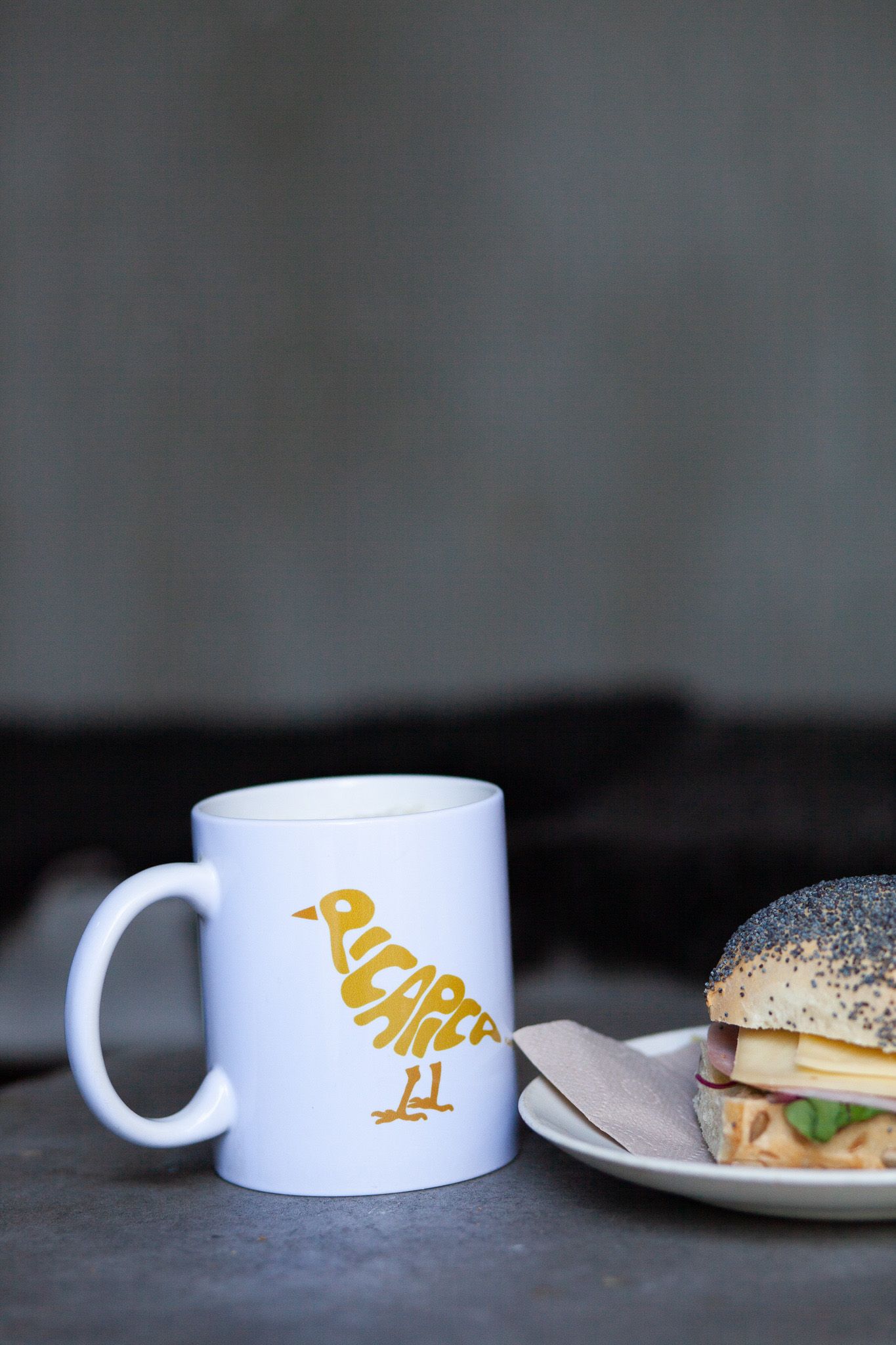 A white coffee mug with a golden bird logo sits beside a sandwich on a plate against a dark background.