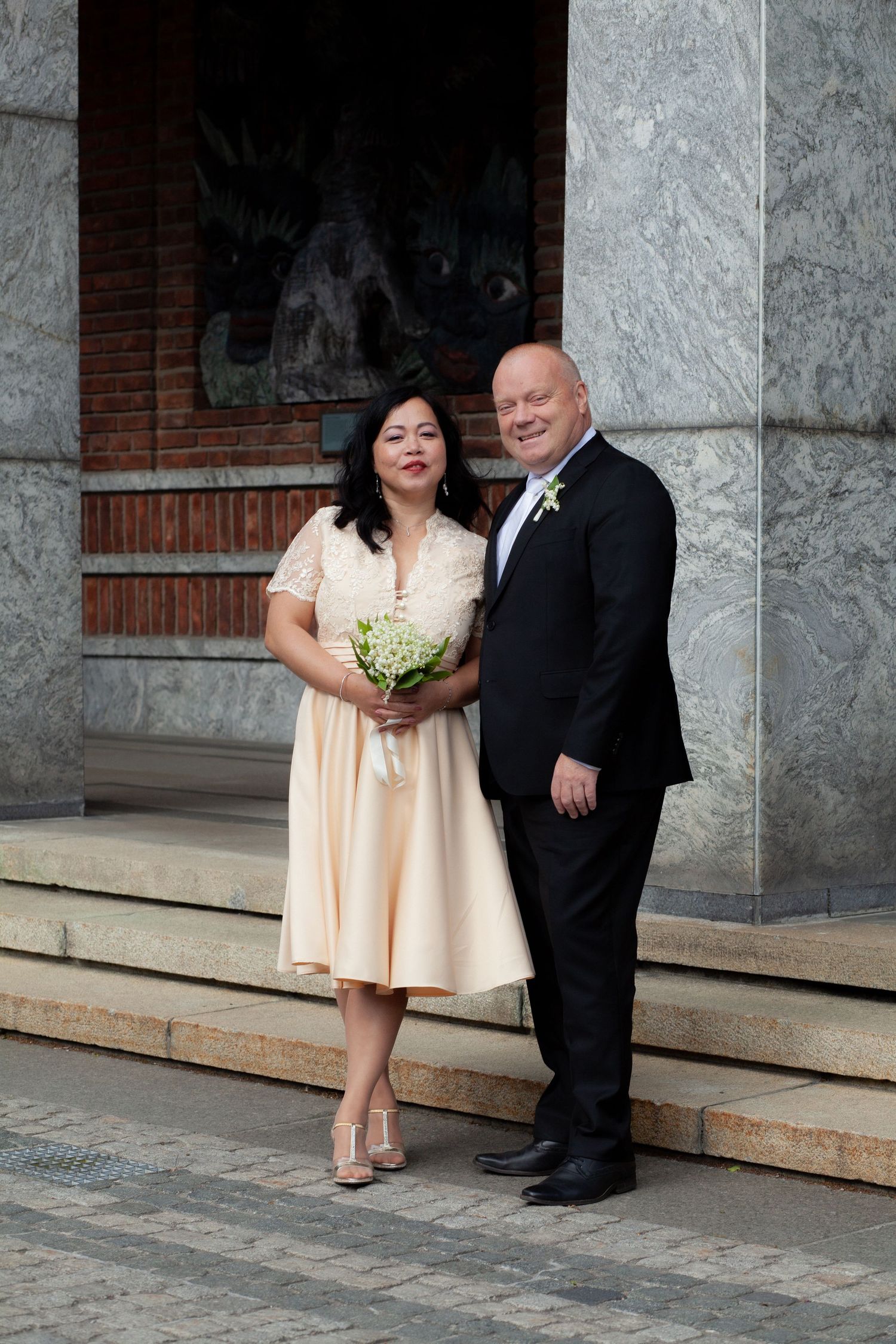 Couple in wedding attire posing on steps, woman in peach dress holding bouquet, man in black suit with boutonniere.