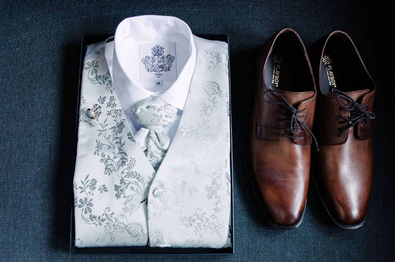 Wedding attire details featuring white dress shirt and brown leather shoes on dark surface.