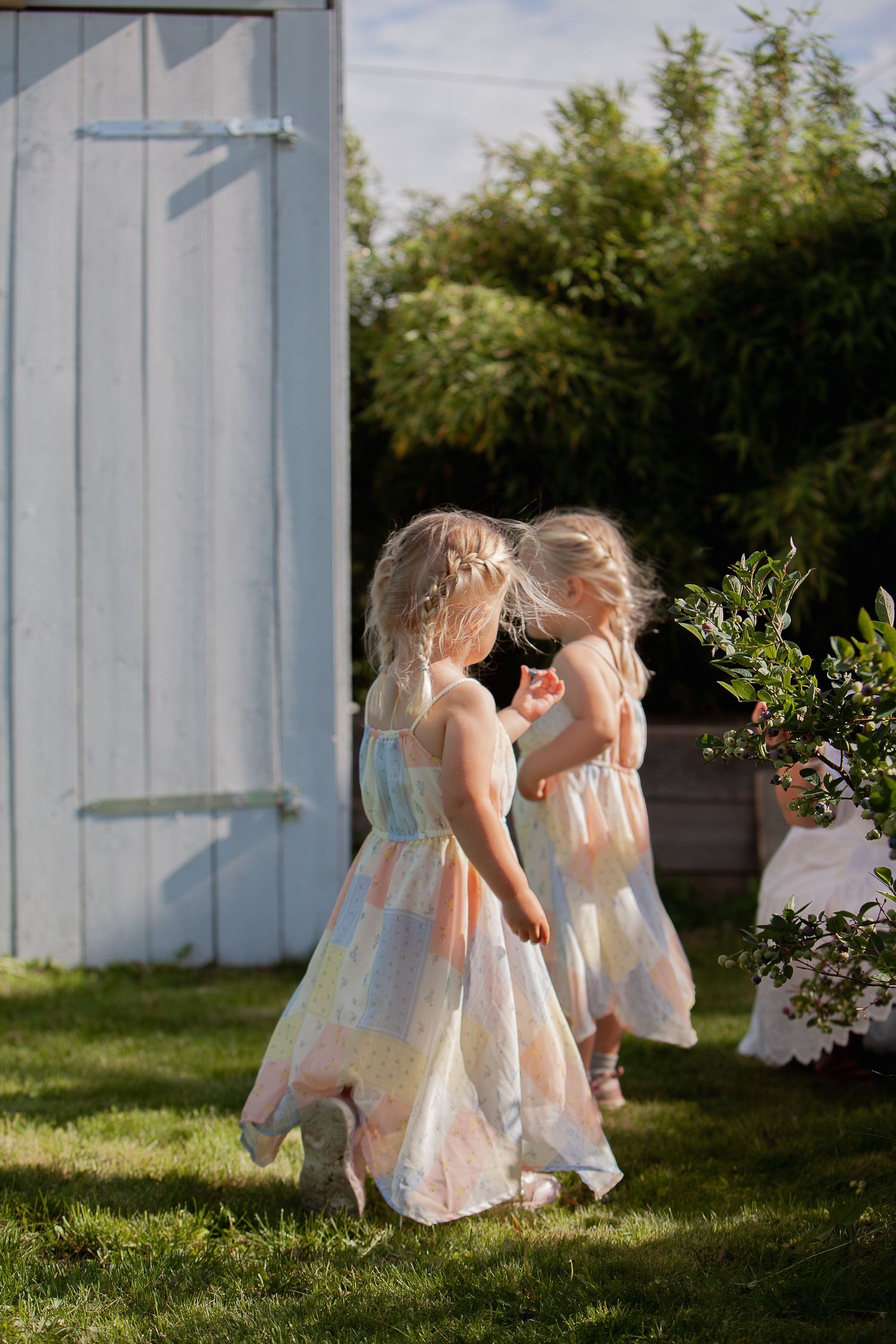 Two children in matching light dresses walking together near a white garden shed.