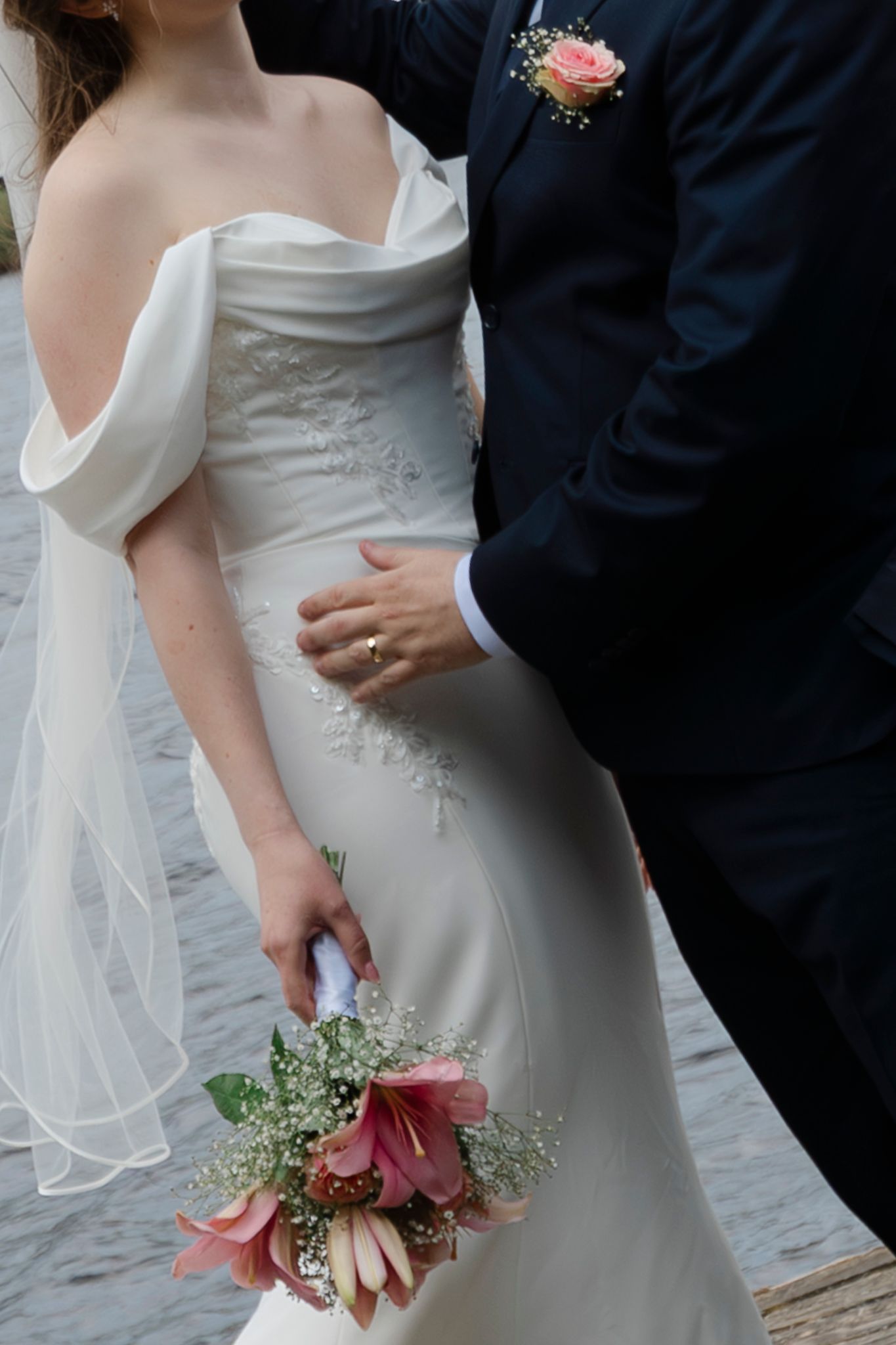 Bride in off-shoulder white gown holds pink bouquet while groom in navy suit with boutonniere embraces her waist.