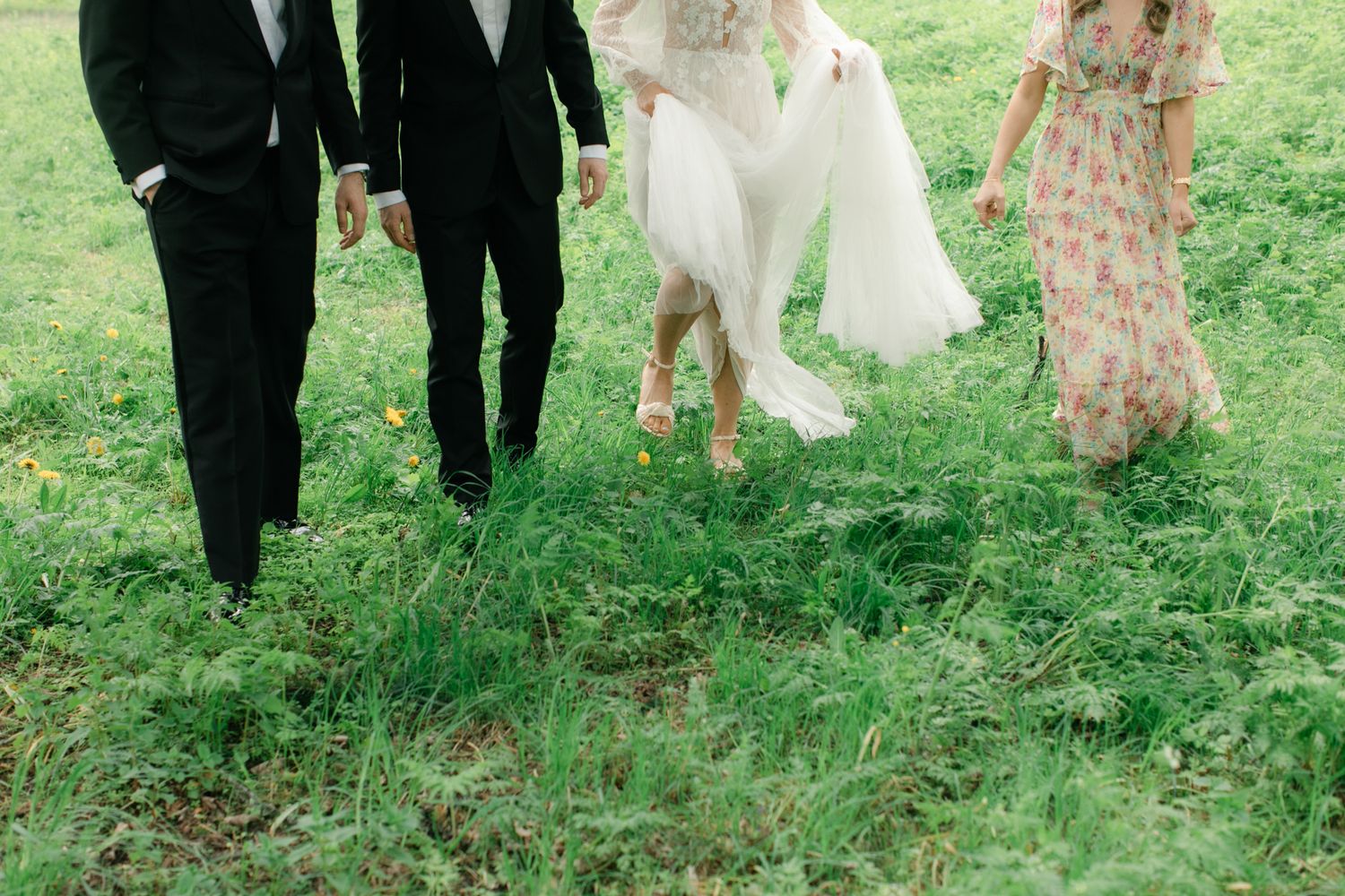 Wedding party walking through lush green grass, bride in white gown and bridesmaid in floral dress.
