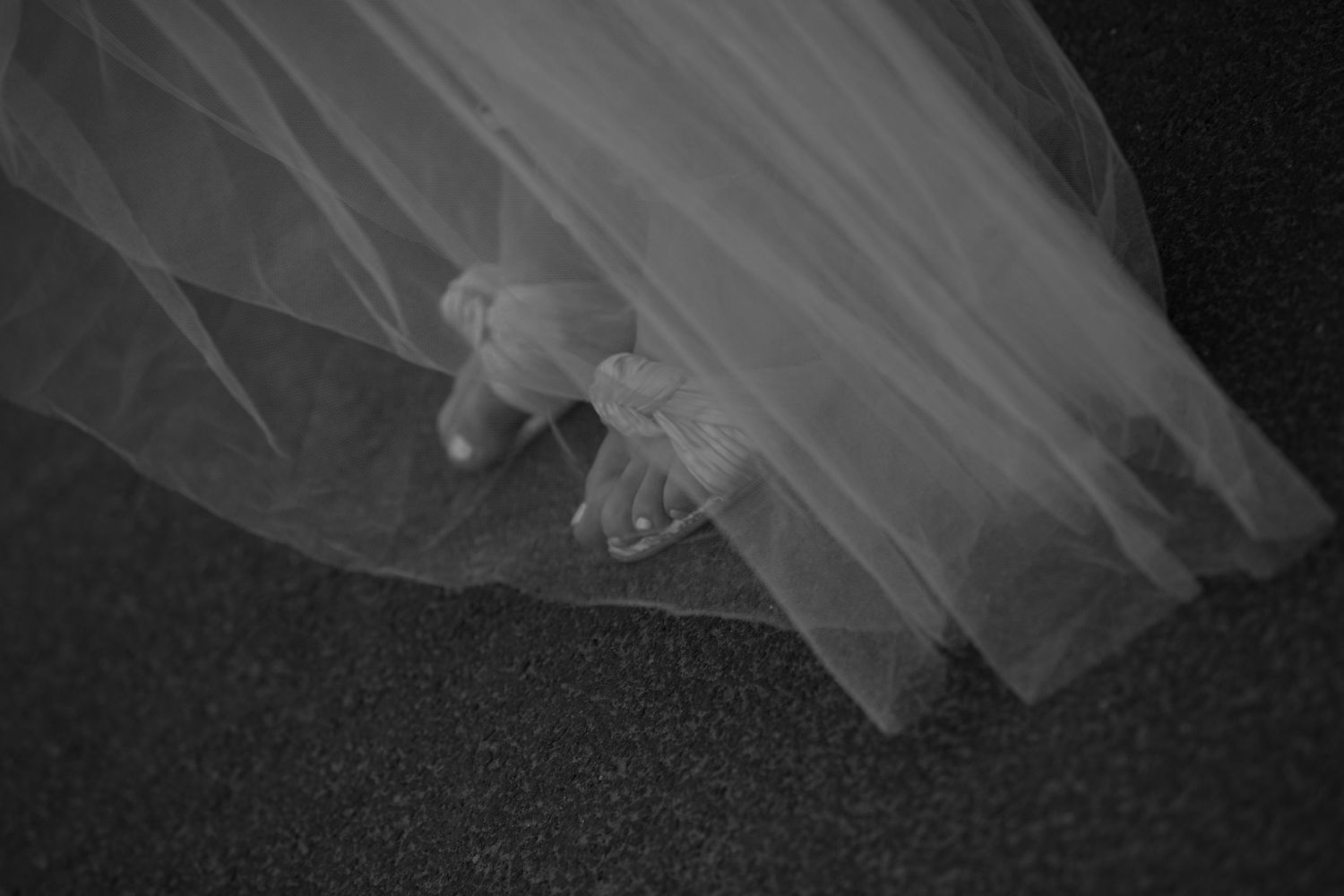 Black and white photo of a wedding dress veil draped over white bridal shoes on a dark floor.