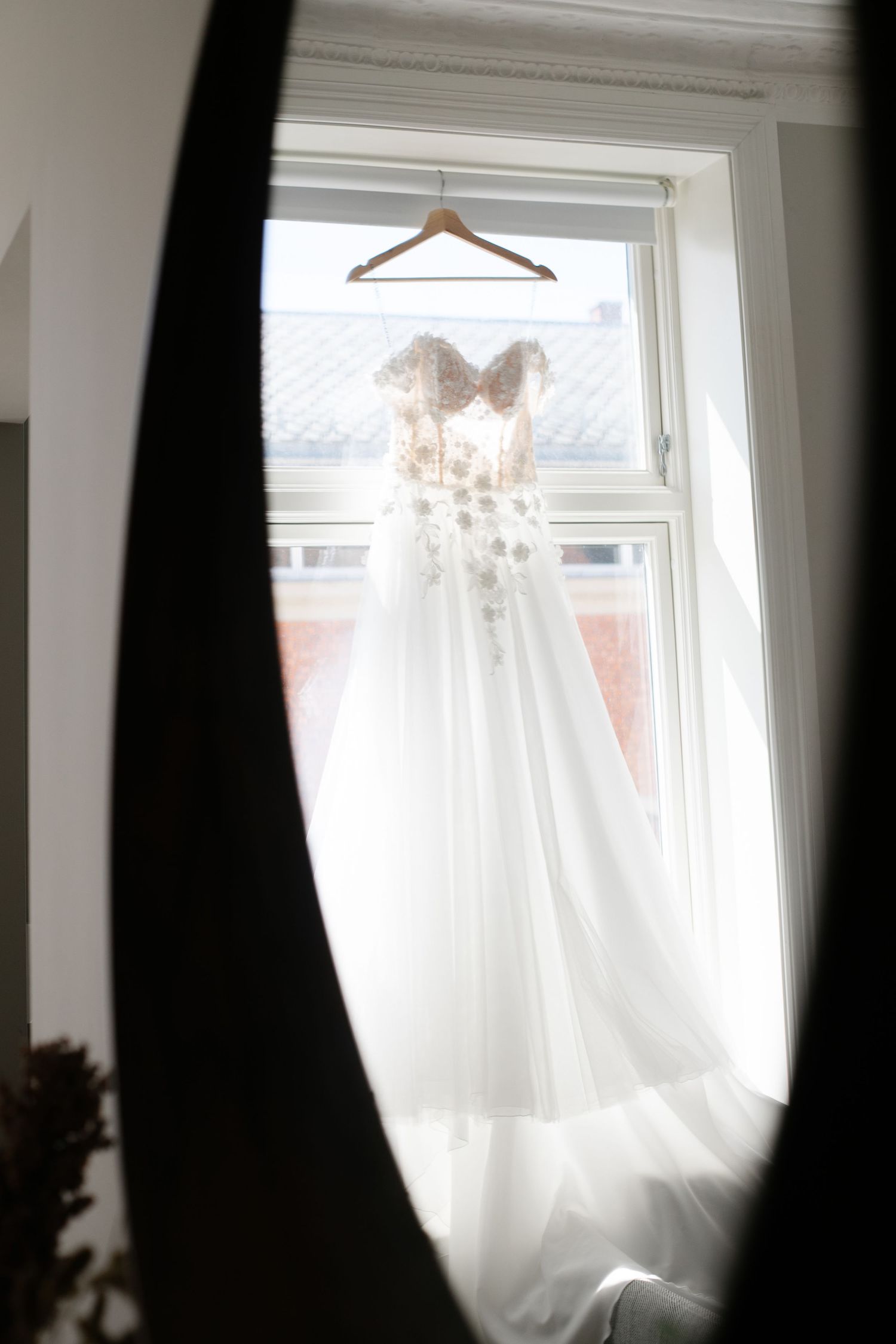 A delicate white wedding dress with lace details hangs in a sunlit window on a wooden hanger.
