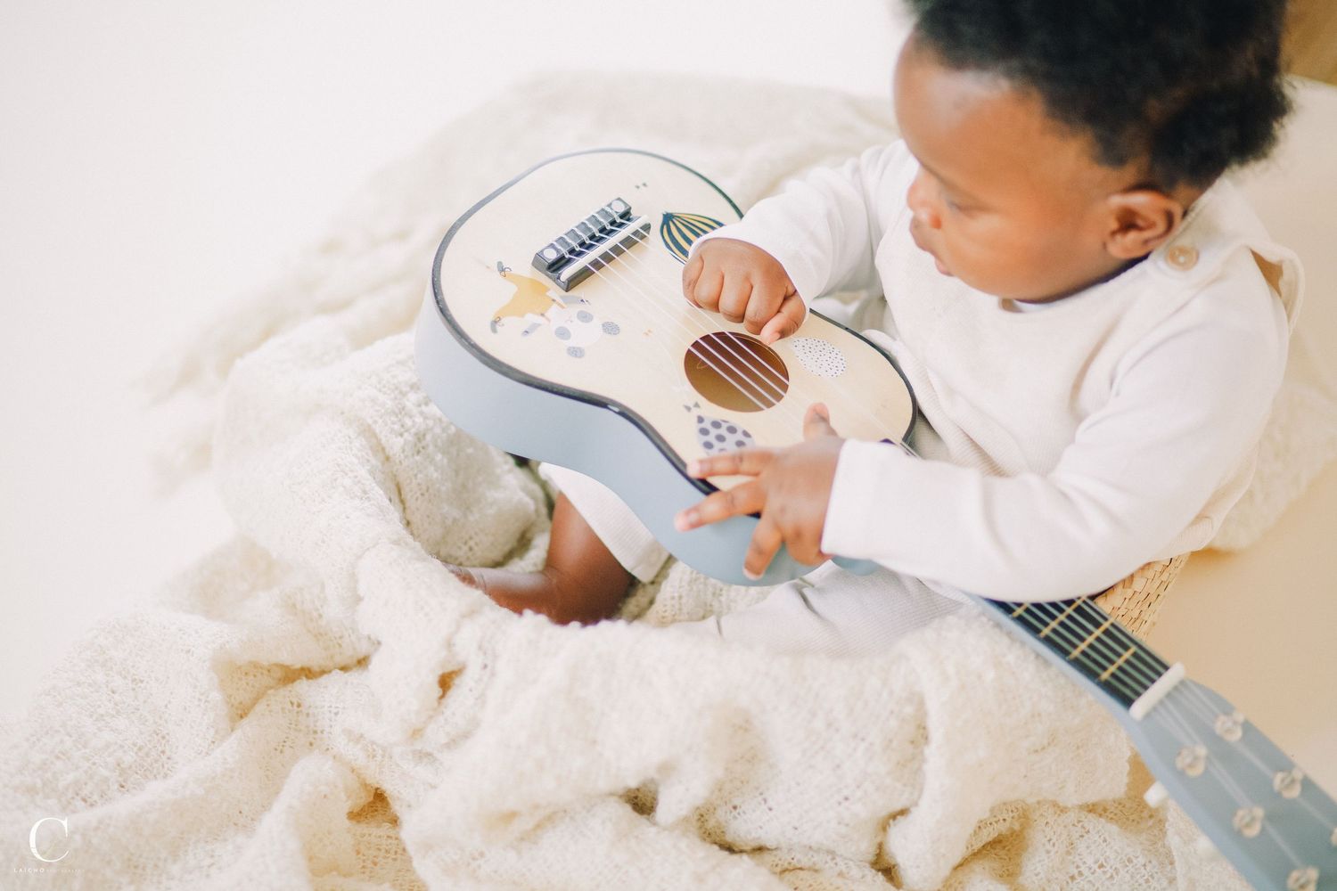 Baby wearing white outfit playing with musical instrument on light background.