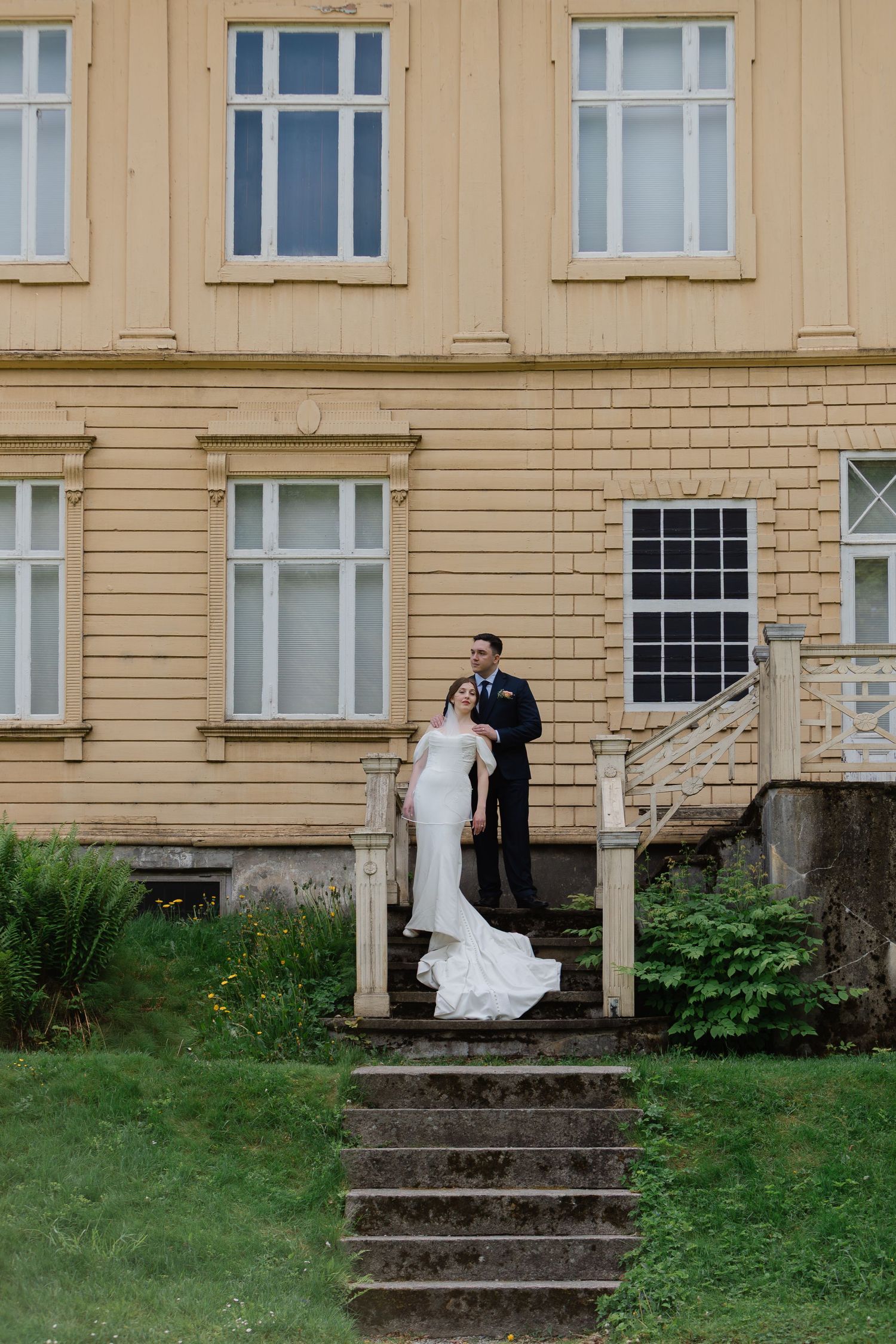 Bride and groom stand on stone steps of yellow manor house in elegant wedding attire.