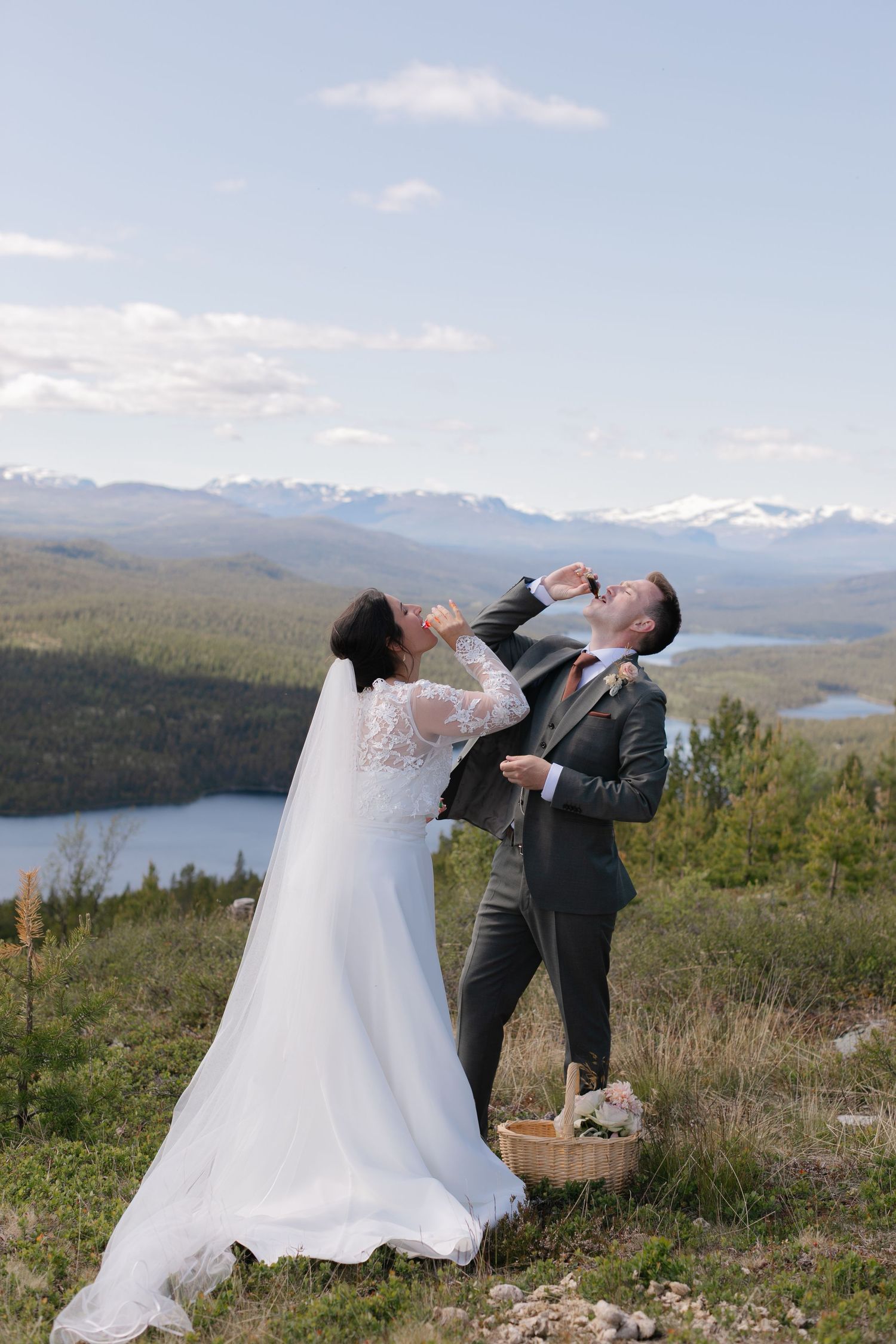 A playful wedding couple poses dramatically on a hilltop with mountains and lake in the background.