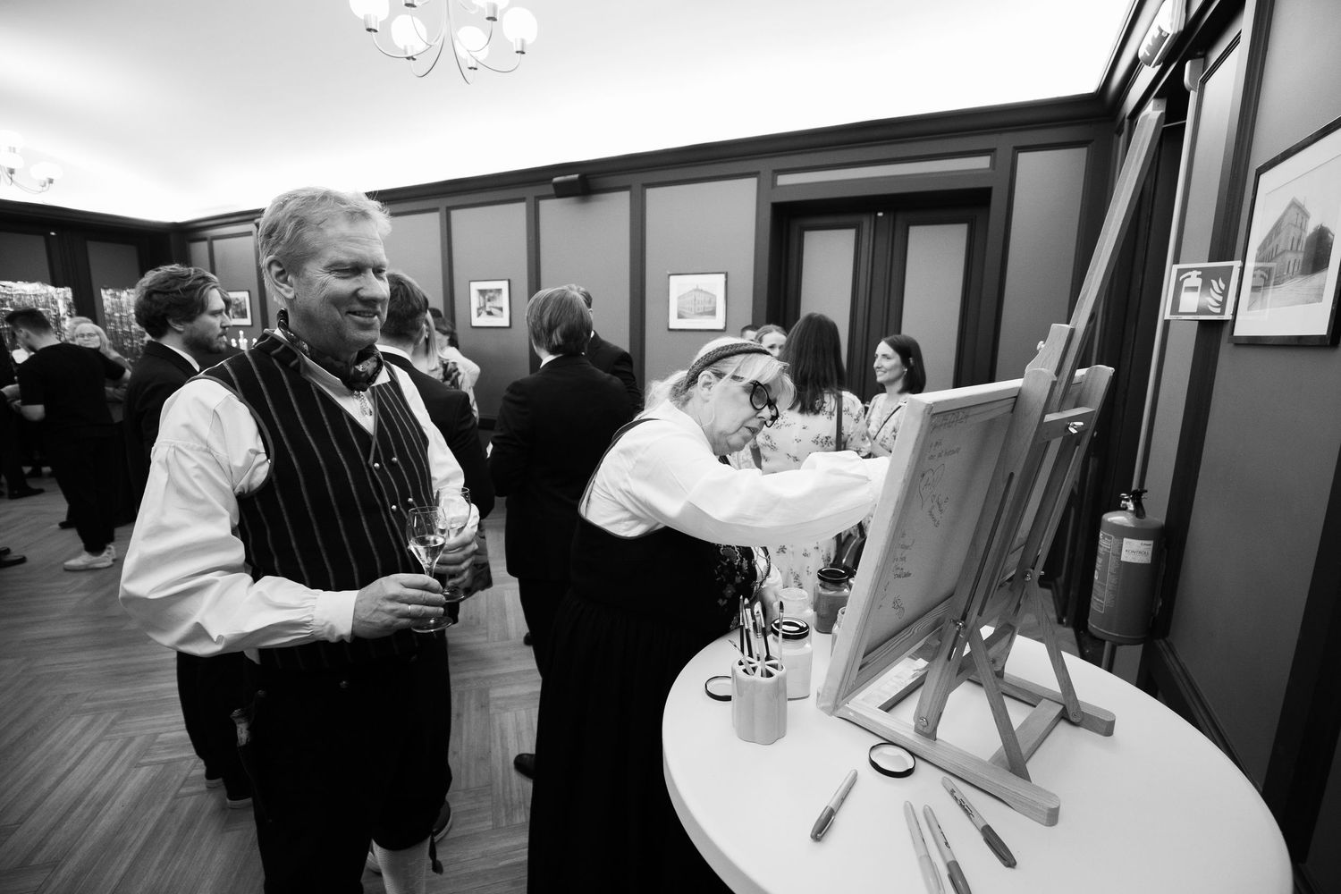 People in traditional formal attire gather around an easel at an indoor event.