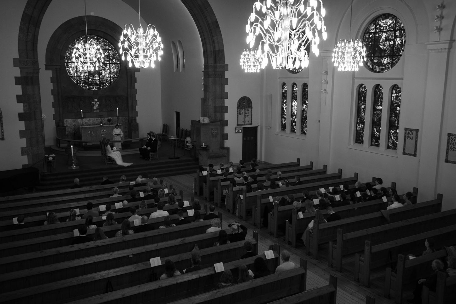 Black and white photo of a church interior with ornate chandeliers, stained glass windows, and rows of wooden pews filled with people.