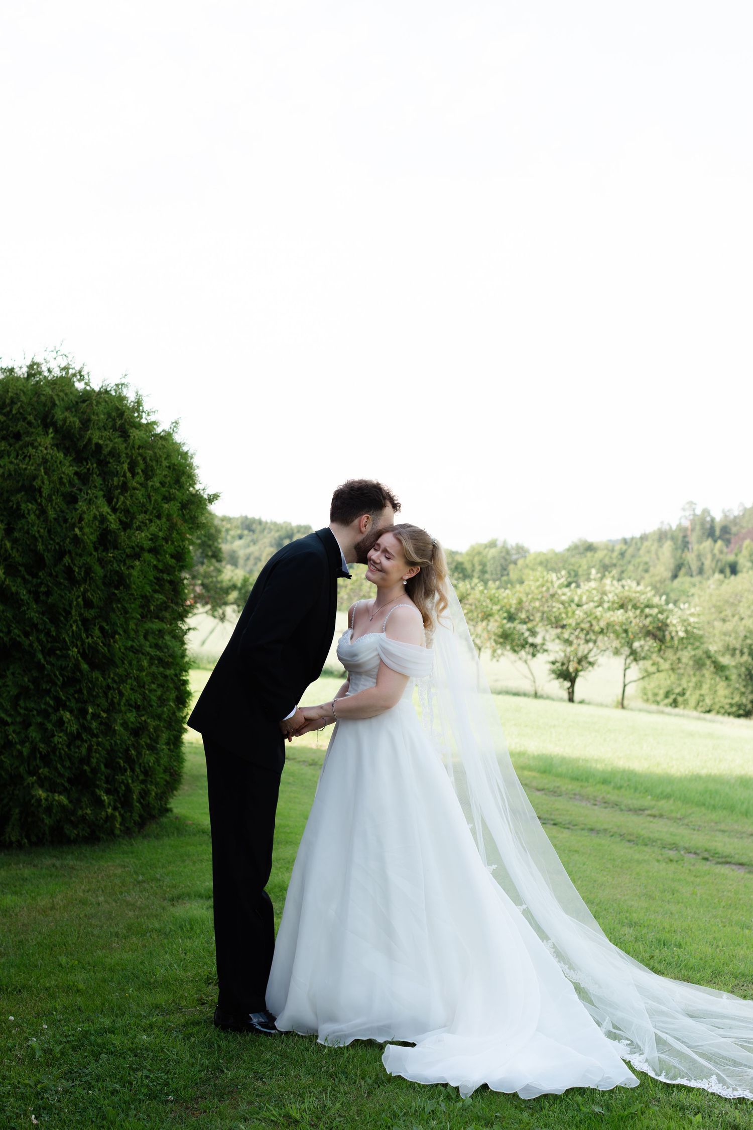 Bride and groom share a romantic moment outdoors on lush green grass, with rolling hills in the background.