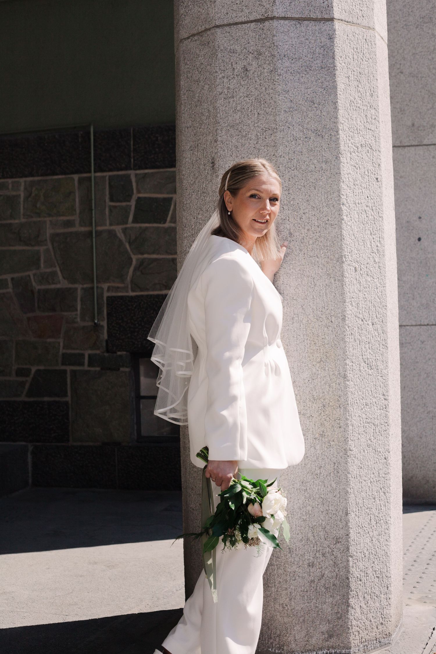 A bride in white poses elegantly between stone columns in natural sunlight.
