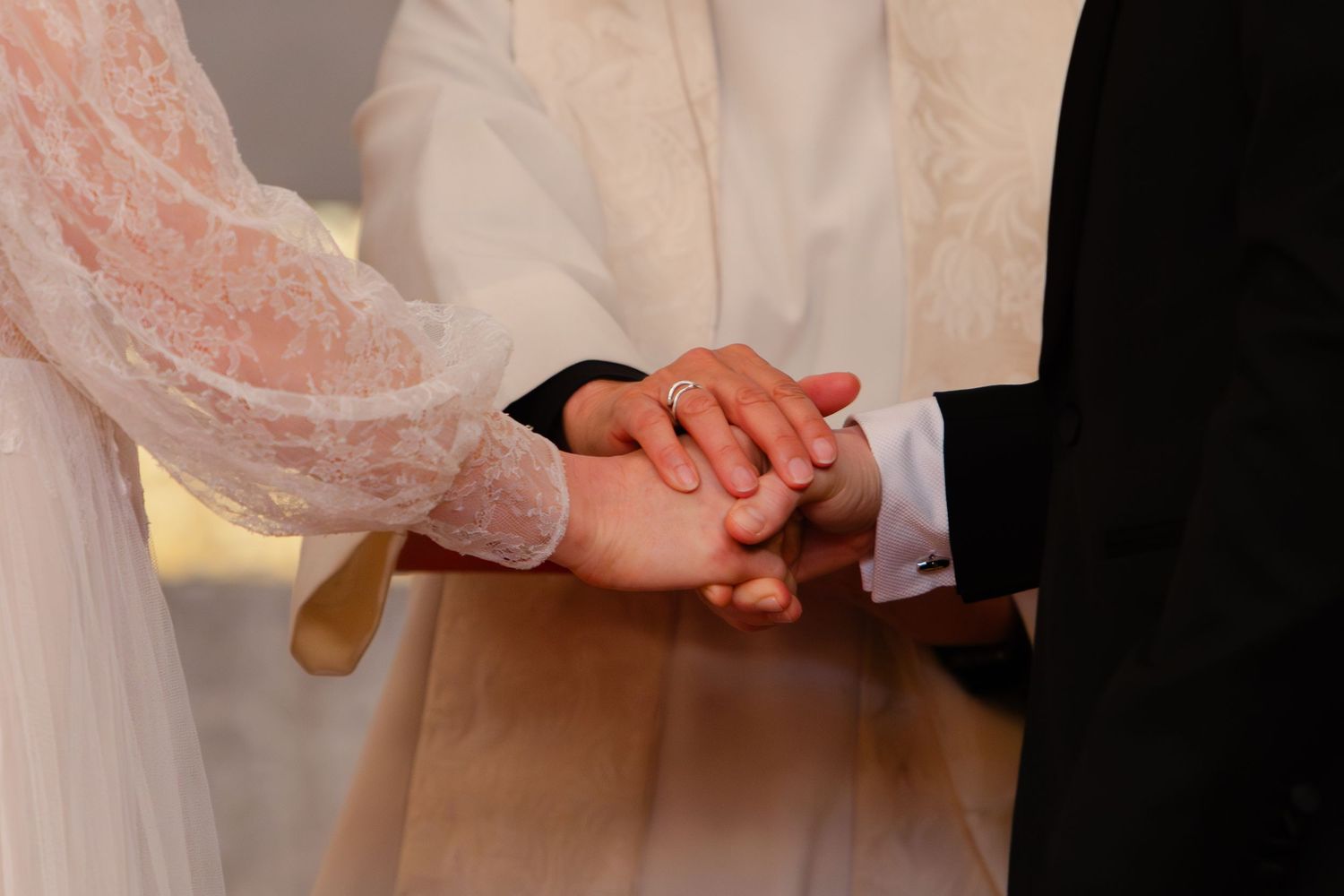 Bride and groom holding hands during wedding ceremony, bride wearing white lace dress with officiant in background.