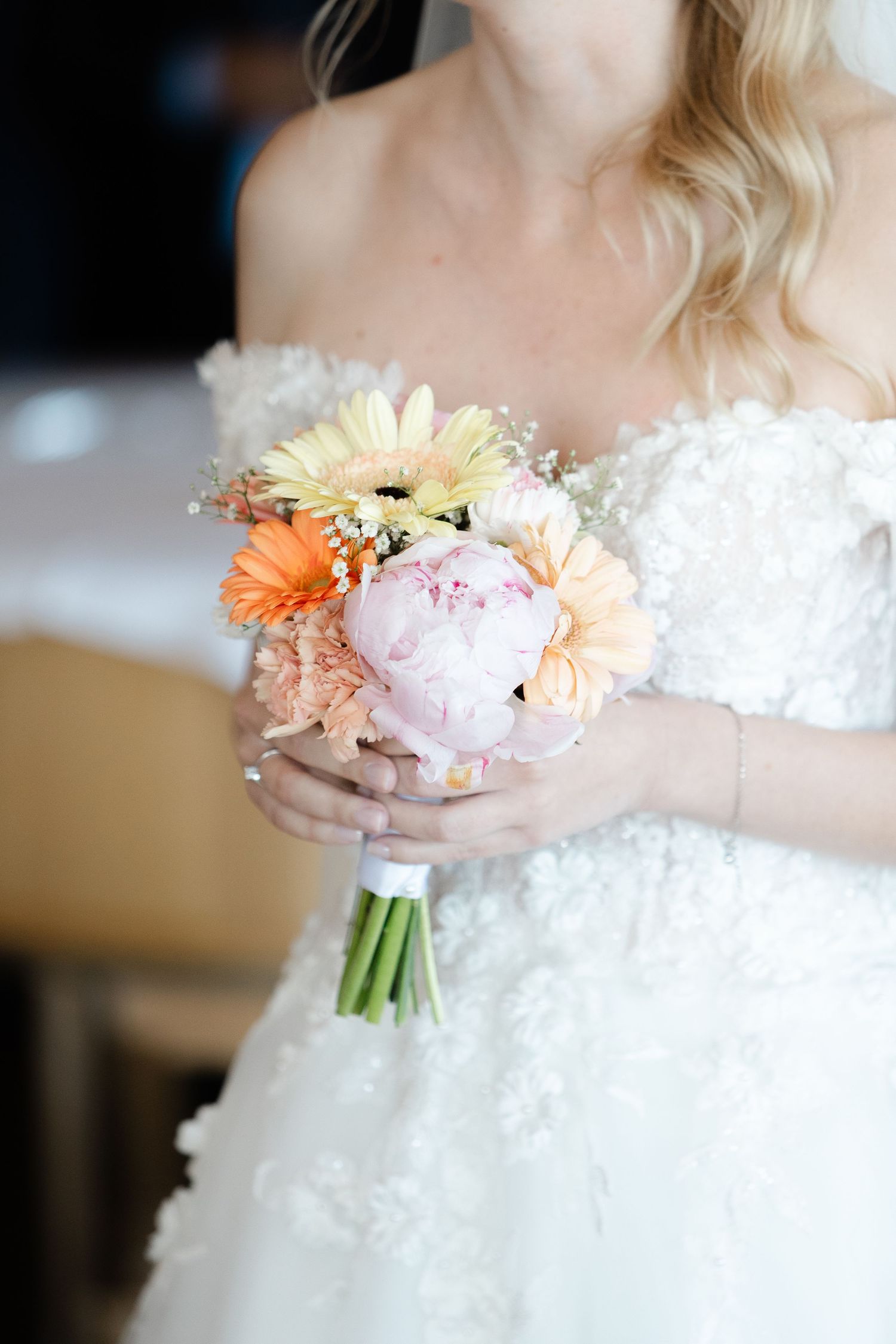 A delicate bridal bouquet with pink peonies, peach gerberas and white baby's breath held against a white lace wedding dress.