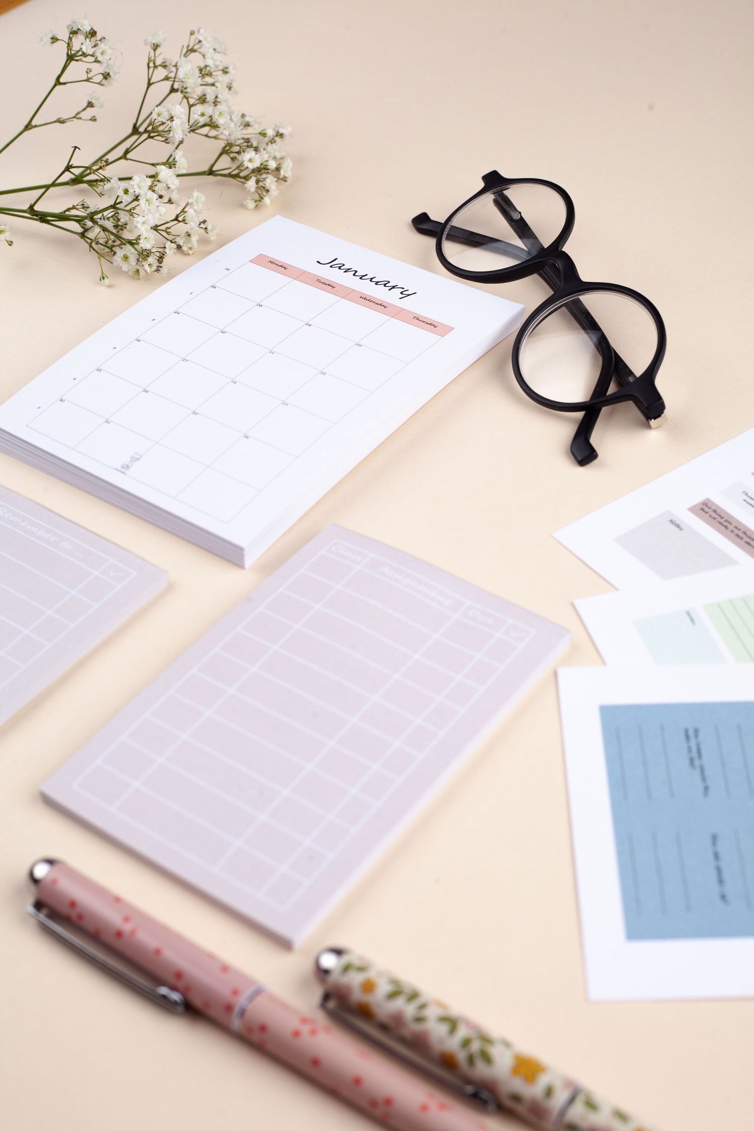 A minimalist desk setup with calendar pages, black round glasses, delicate white flowers, and decorative pens on beige surface.