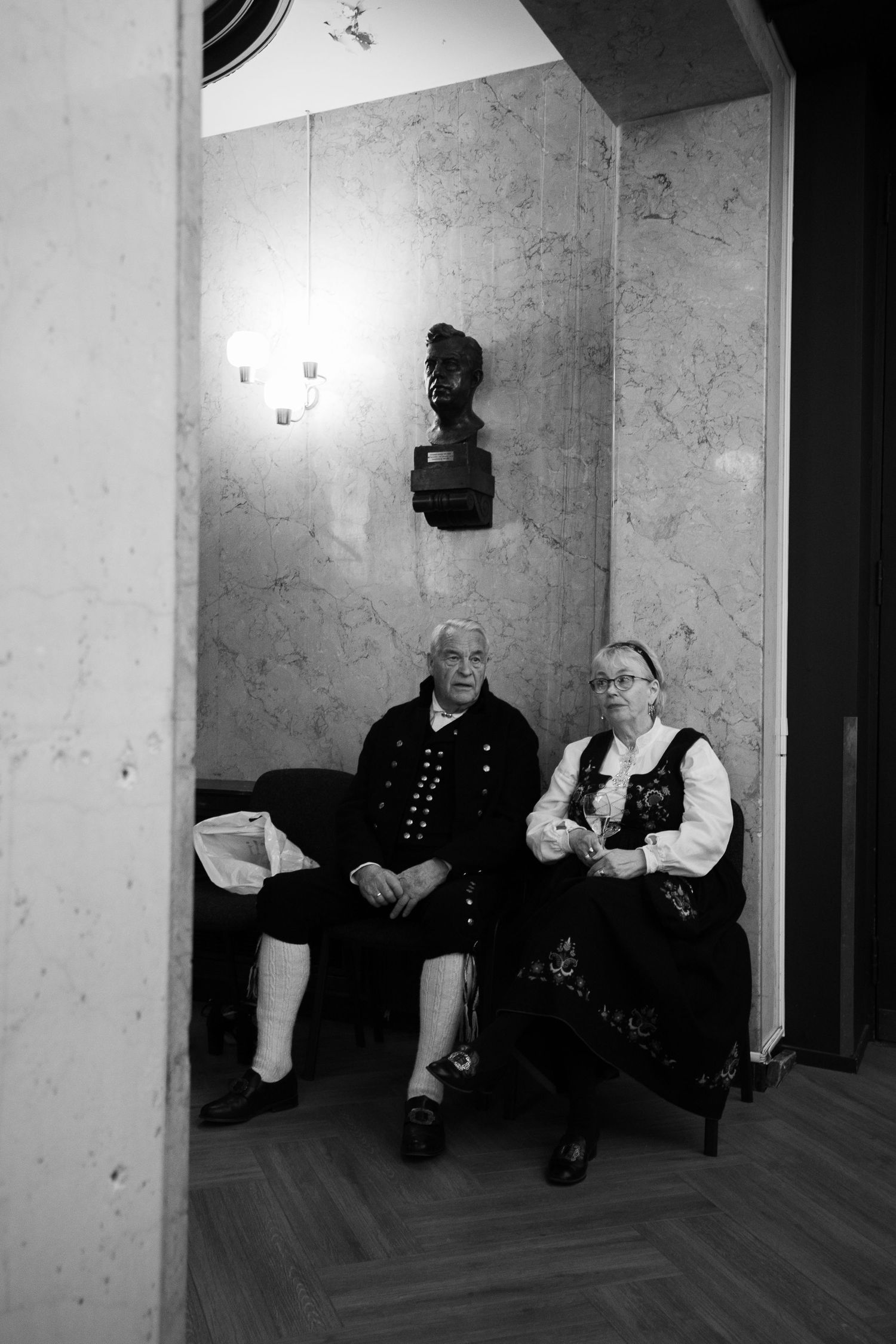 Two people in traditional dress sit beneath a decorative wall bust in a hallway.