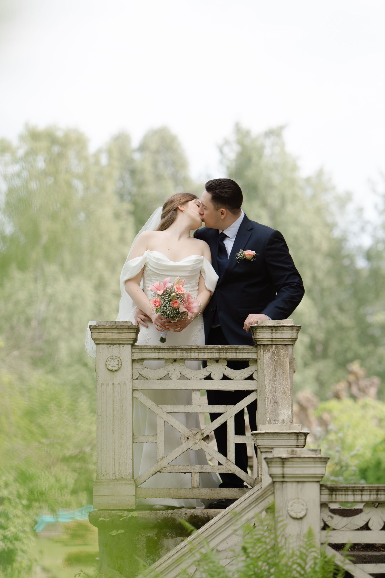 Bride and groom share a romantic kiss on a stone bridge staircase, bride holding pink bouquet, lush green trees background.