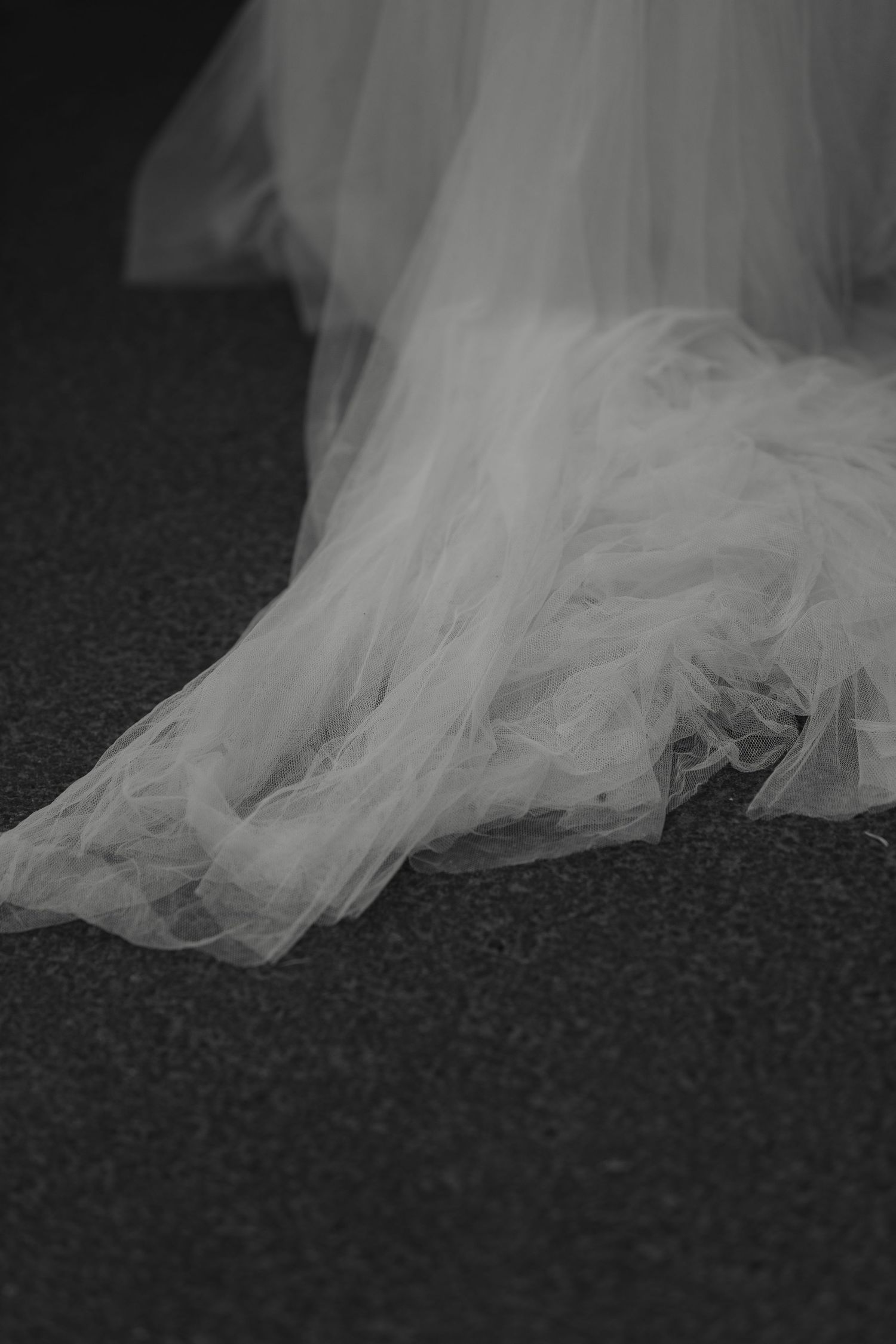 Black and white photo of a flowing tulle wedding dress train spread elegantly on a dark floor.