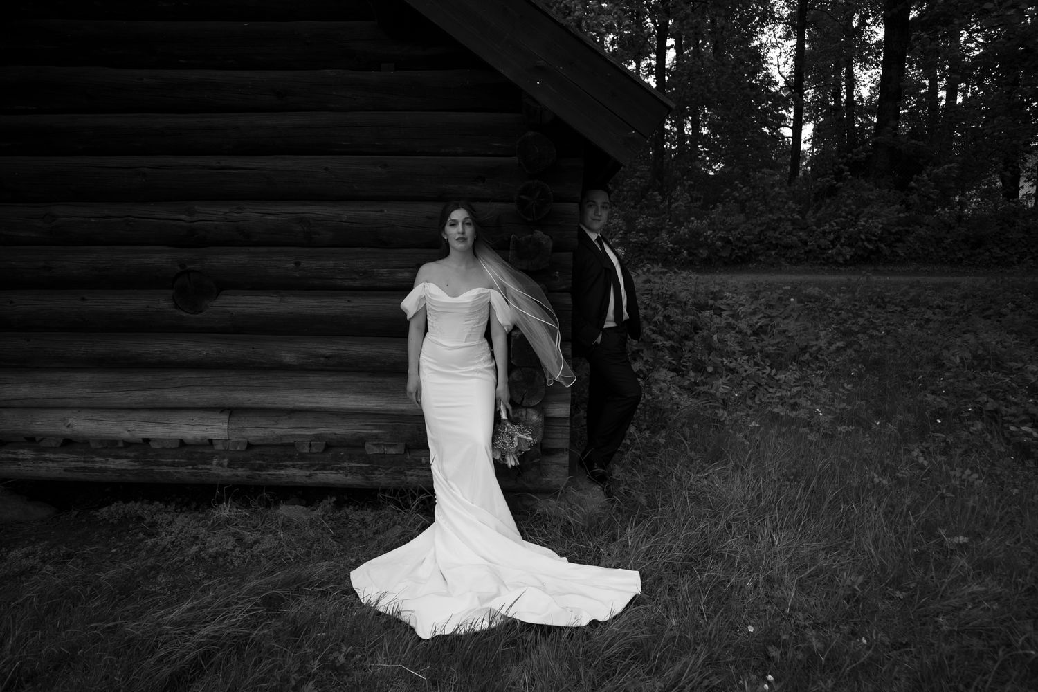 Black and white wedding photo of bride in fitted gown and groom walking beside a rustic log cabin in a forest.