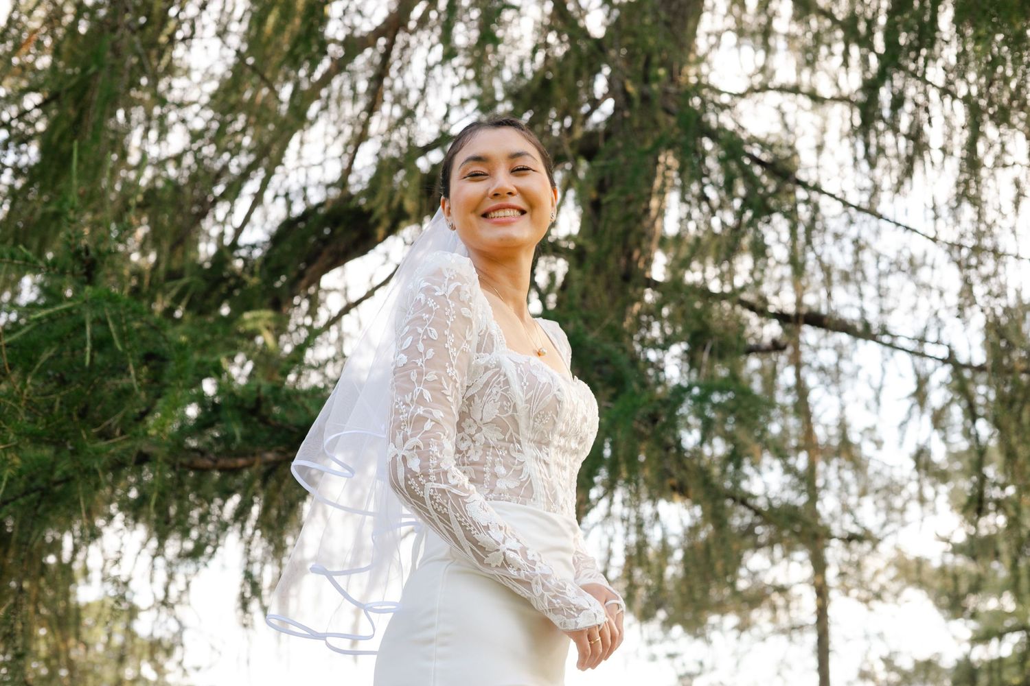 A joyful couple in wedding attire laughs together beneath pine trees.