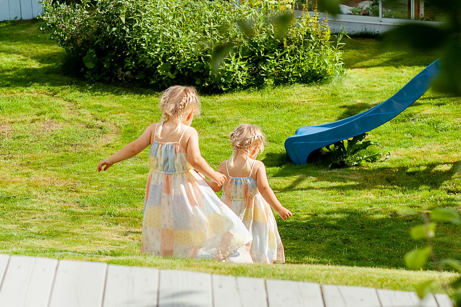 Children in flowing dresses playing in a sunny garden with a blue slide.