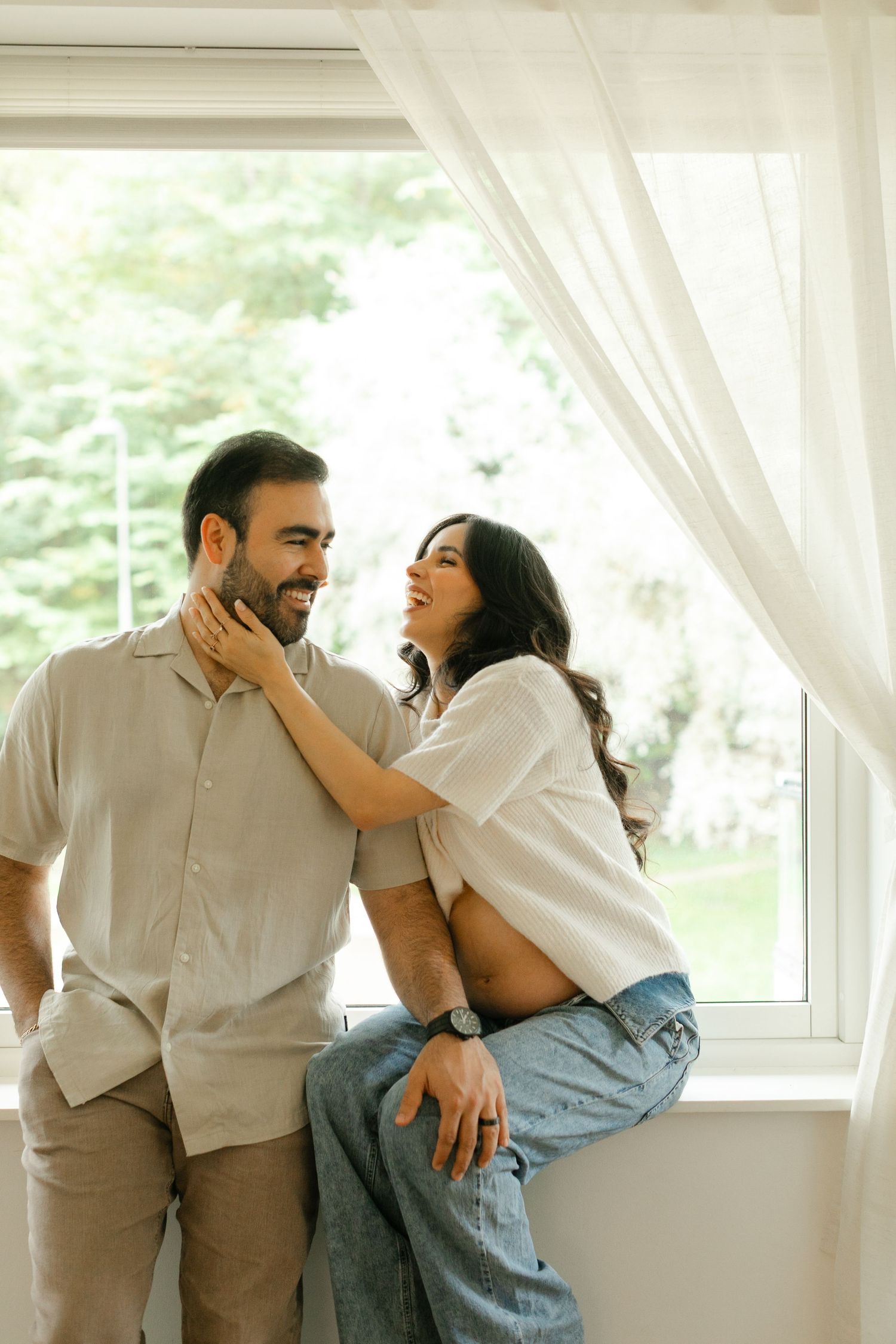A couple shares intimate laughs and playful moments in a bright sunlit room with white curtains.