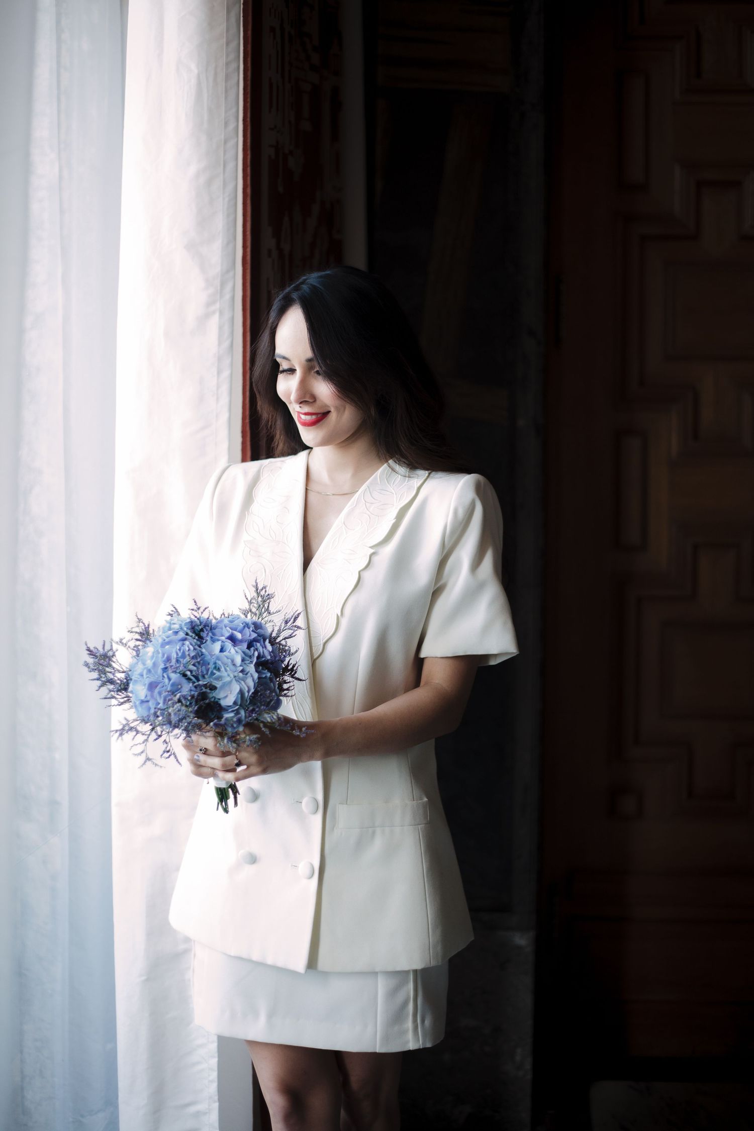 Soft window light illuminates a person in white holding a blue floral bouquet.