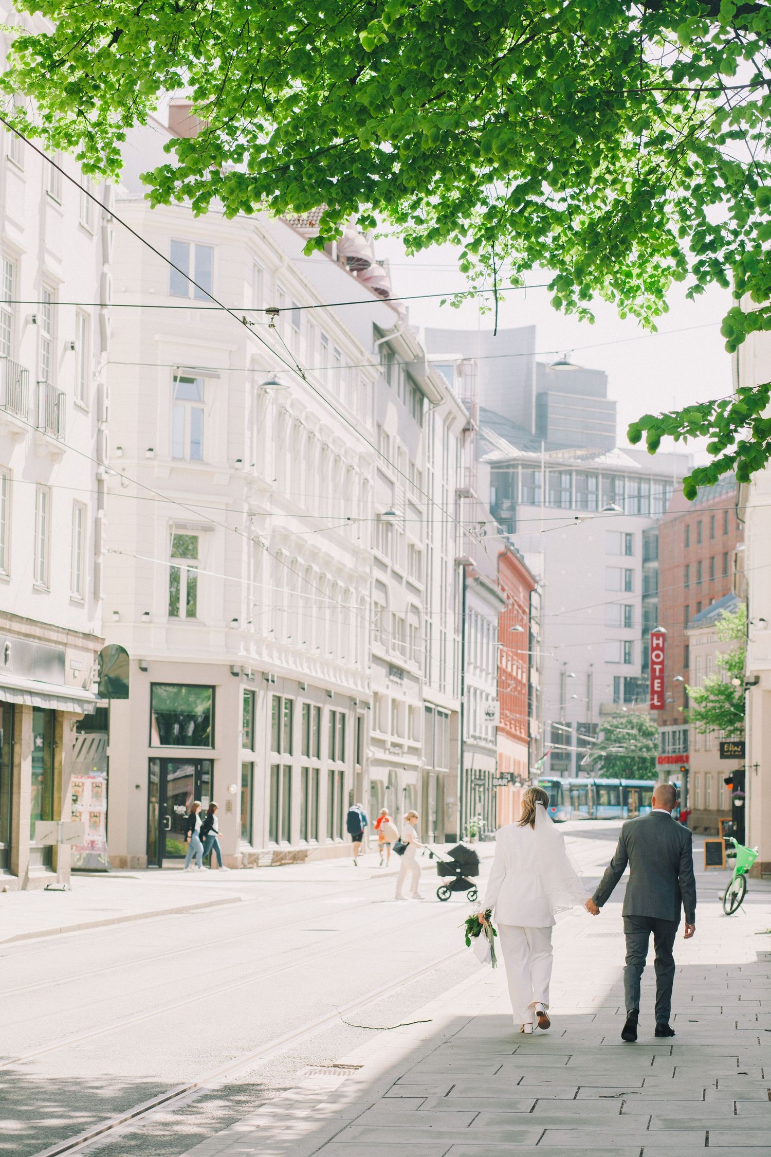 A tree-lined urban street with historic white buildings and pedestrians walking on a bright sunny day.