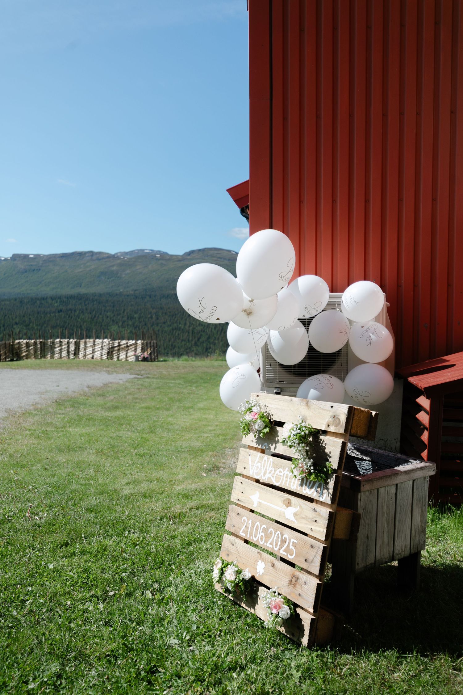 Rustic wooden crates decorated with white balloons sit in a grassy field with mountains in the distance.